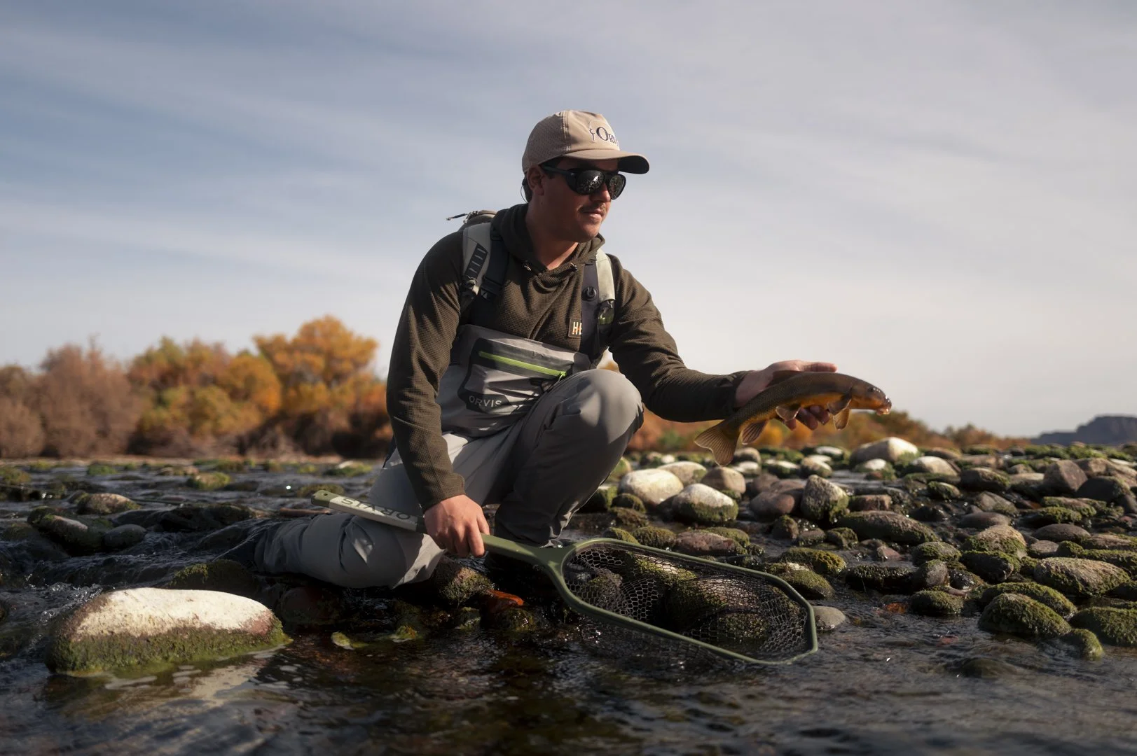 Man kneeling in a stream holding a fish with one hand, with a fishing net in front of him. The man is wearing sunglasses, a beige cap, and outdoor clothing, with autumn trees and a cloudy sky in the background.
