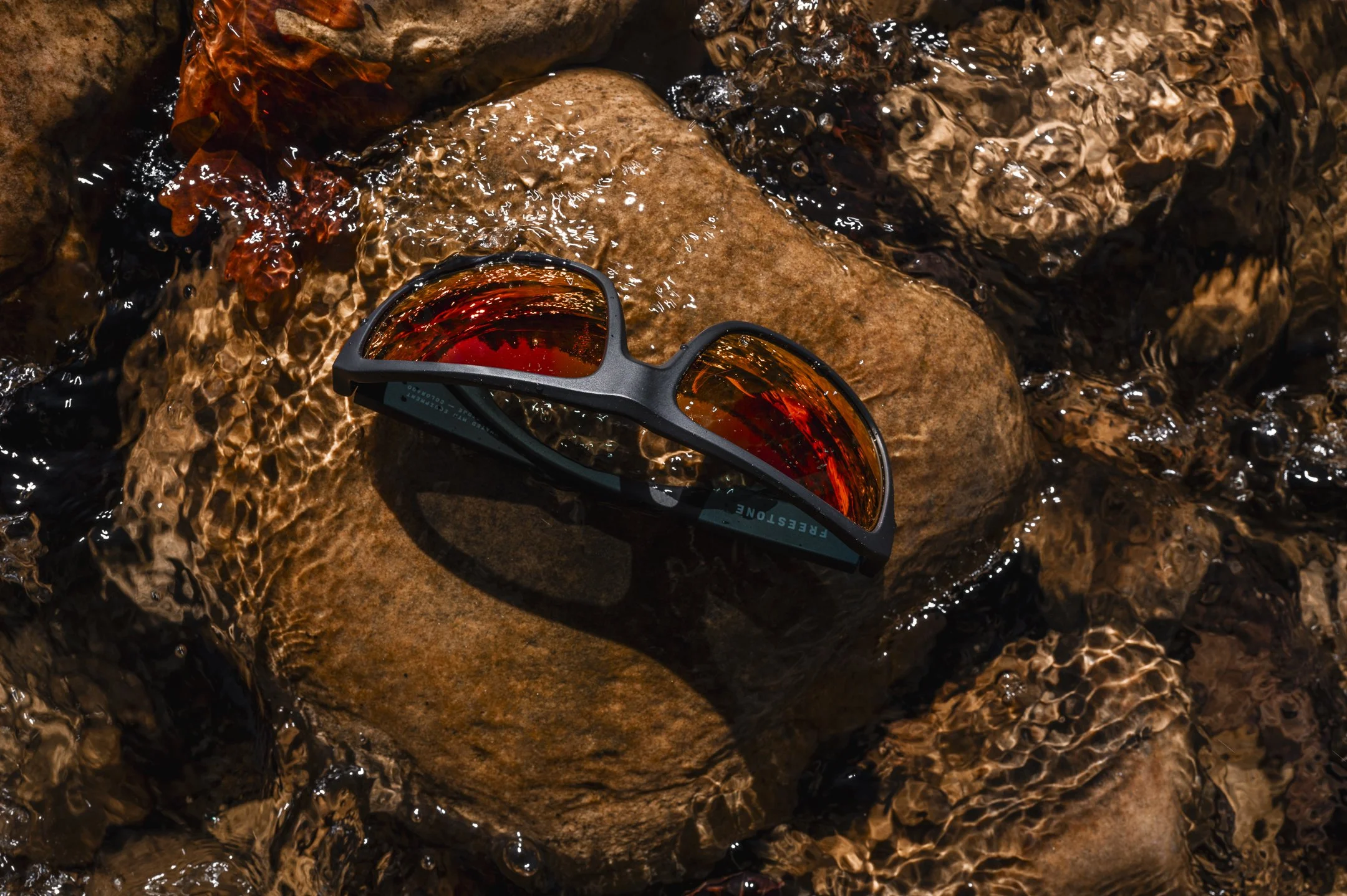 A pair of sunglasses with red-tinted lenses resting on a wet rock in a stream, with clear water flowing around.