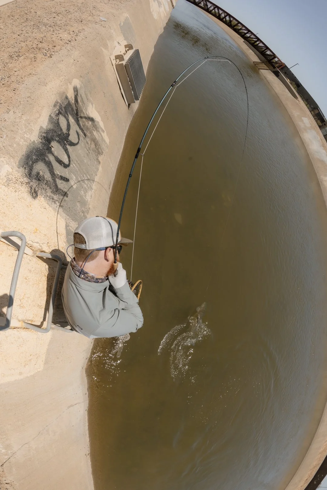 A man fishing by a canal with a bridge in the background, sitting on a concrete ledge, holding a fishing rod, with water splashing from the fish he caught.