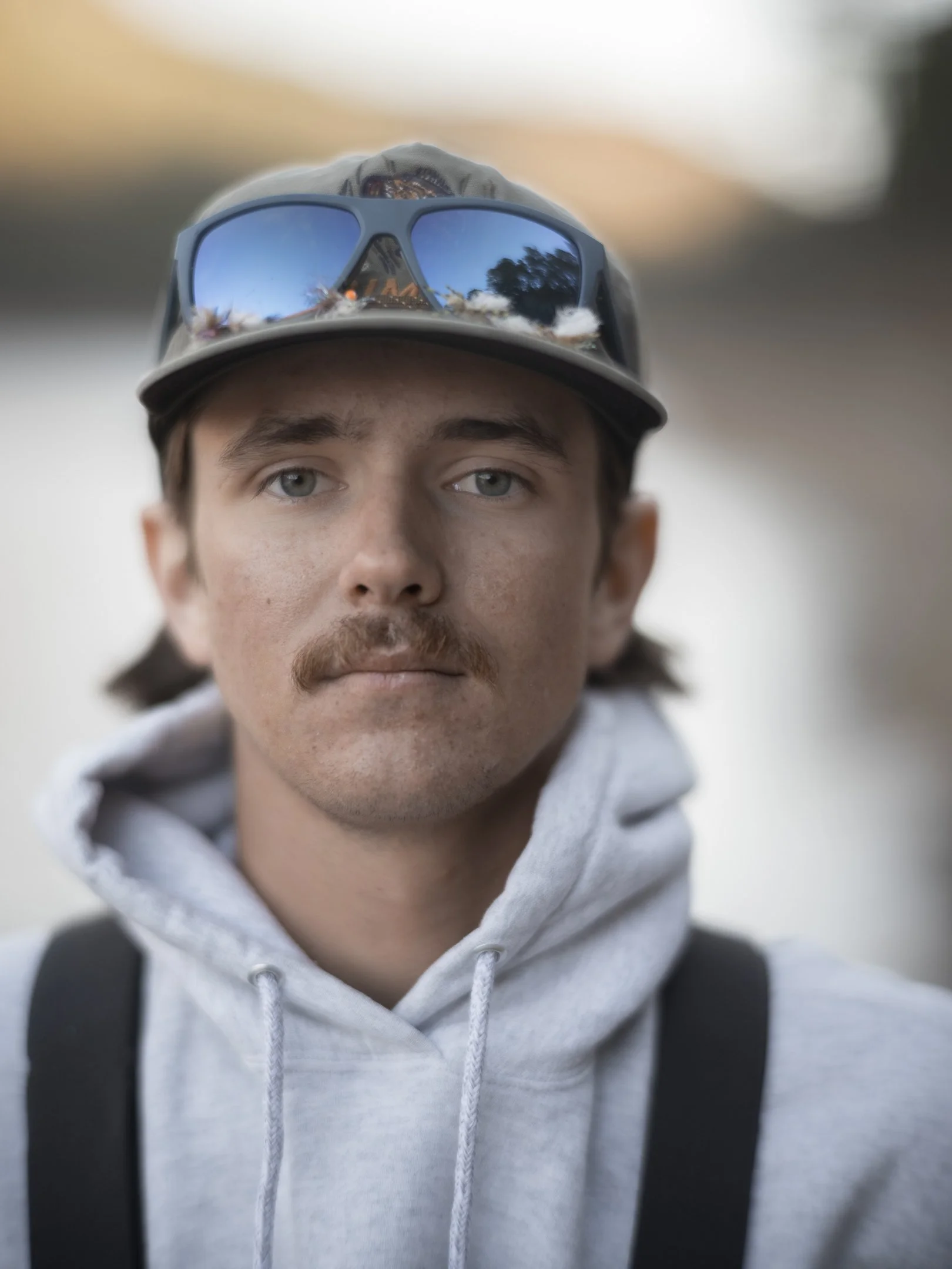 A young man with light skin, brown hair, and a mustache wearing a gray hoodie, a baseball cap, and sunglasses with a reflection of trees and sky in the lenses.