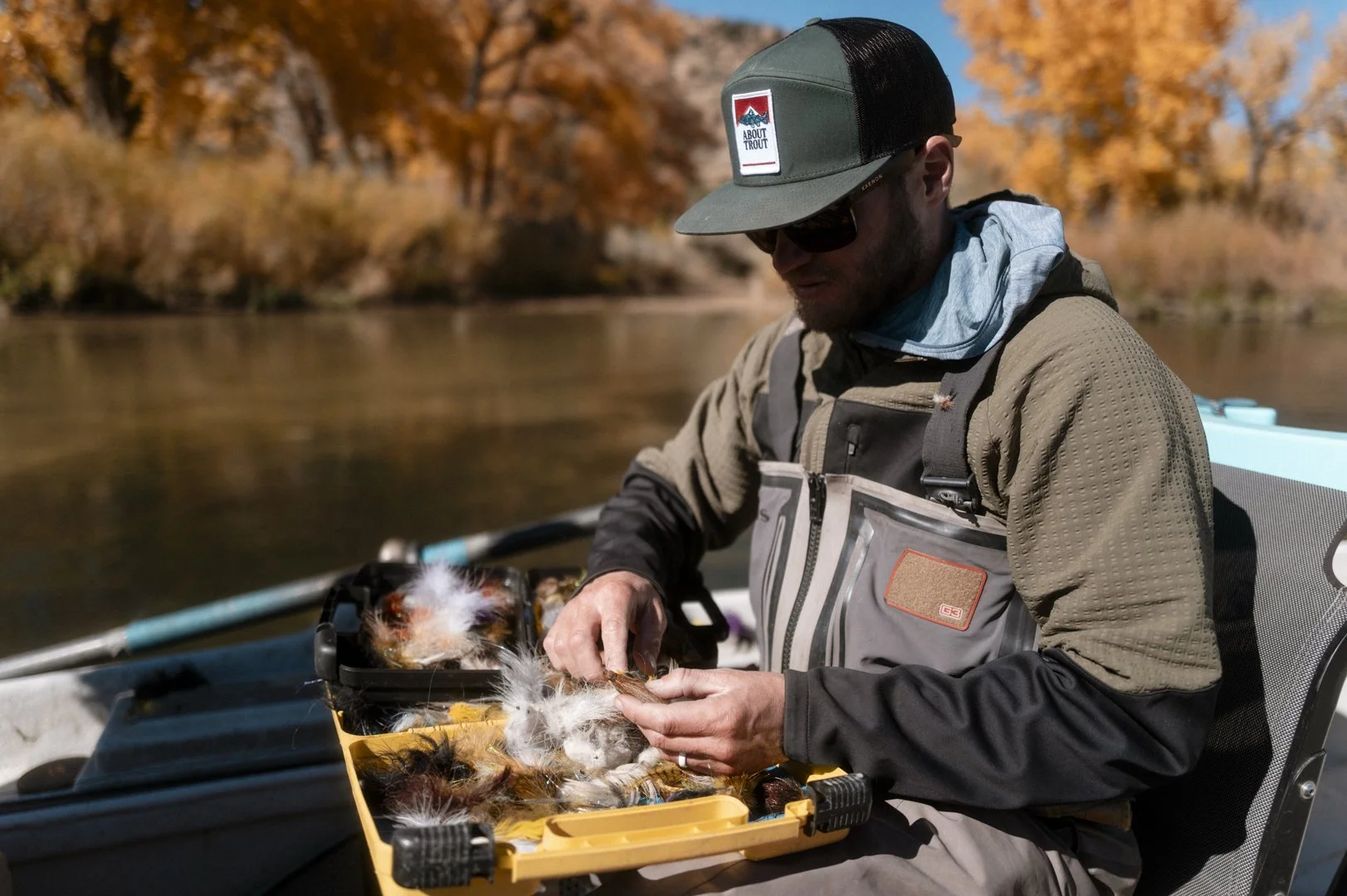 Man in outdoor gear sitting in a boat sorting through a tackle box of feathers and fishing lures during fall with colorful trees in the background.