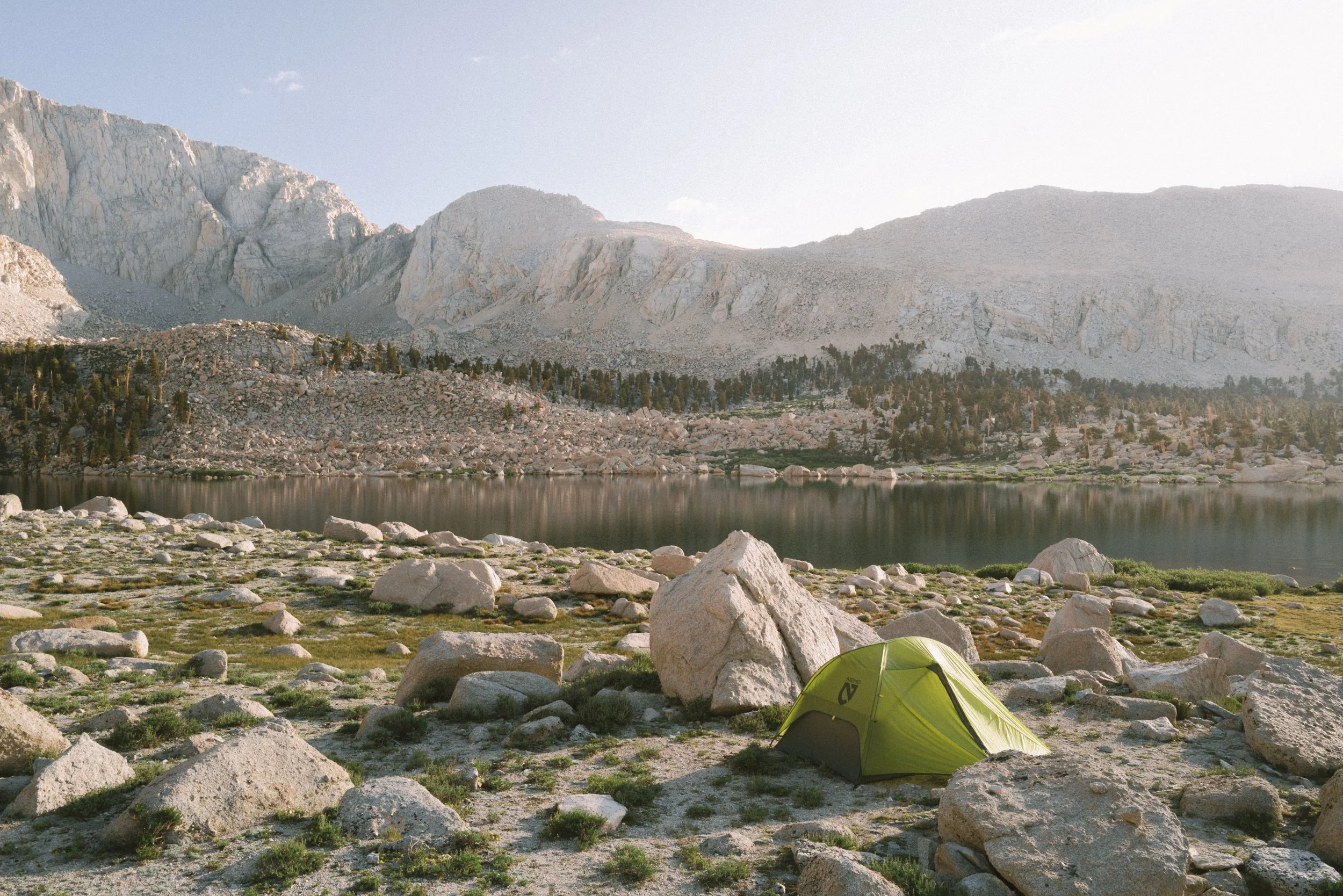 A green tent pitched on rocky, grassy terrain beside a lake with mountains in the background during daylight.