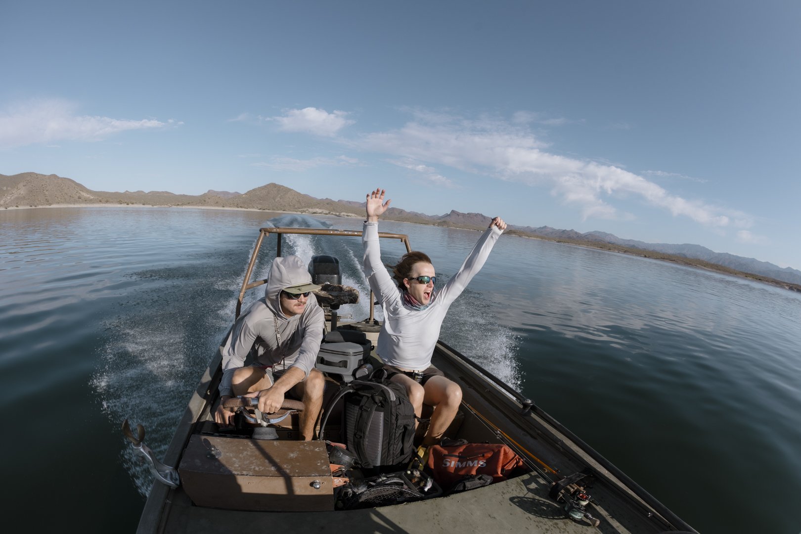 Two people on a boat, one woman with arms raised and smiling, the other man wearing sunglasses and a hoodie, surrounded by lake and mountain scenery.
