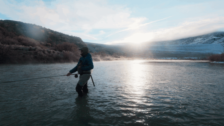 A person fishing in a river during sunset, with distant hills and snow patches in the background.