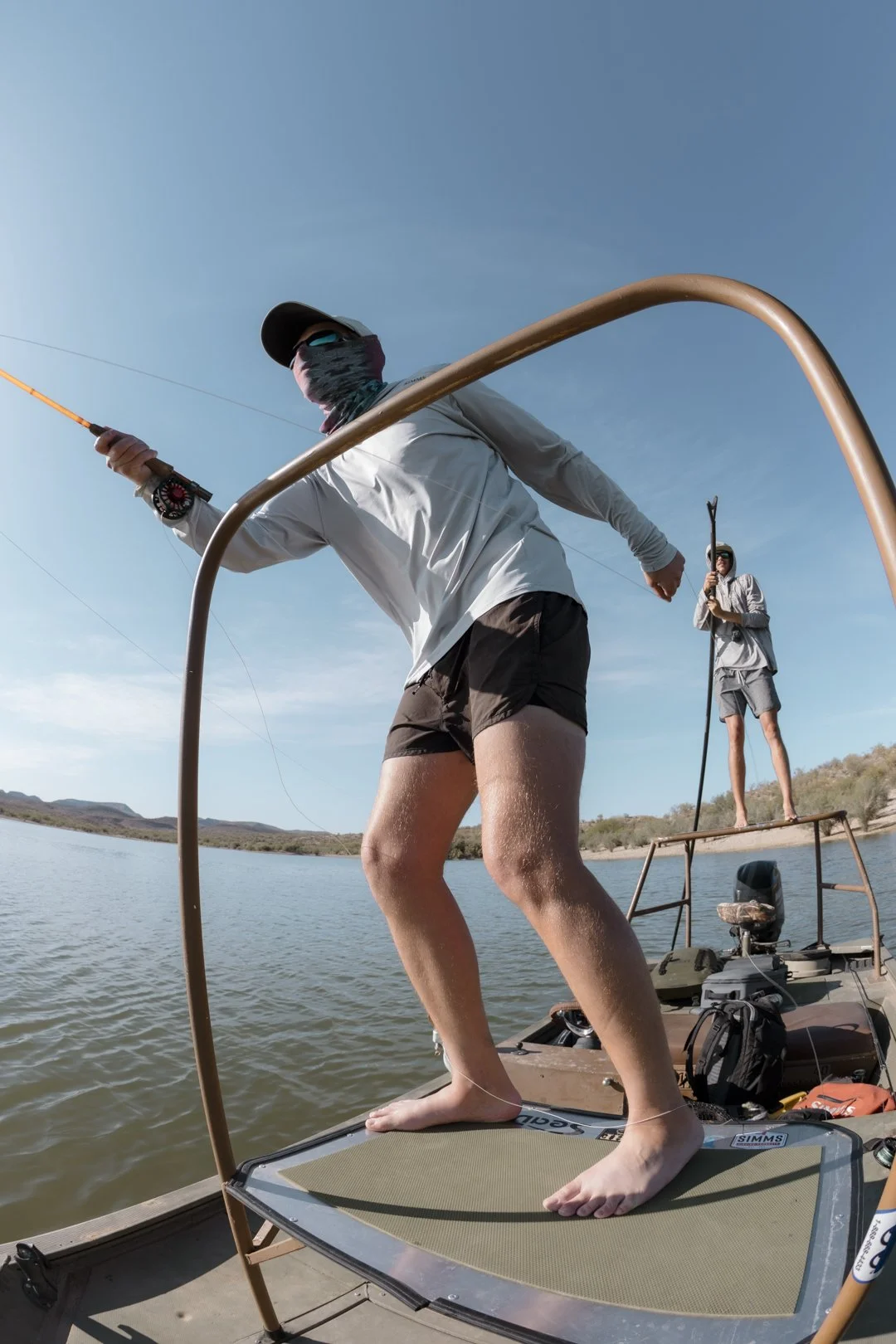 Two people fishing on a small boat with a stand-up paddleboard in a desert lake during daytime. One person is standing on the paddleboard, casting a fishing line, wearing sunglasses, a hat, and a neck gaiter, with bare feet. The other person is stand