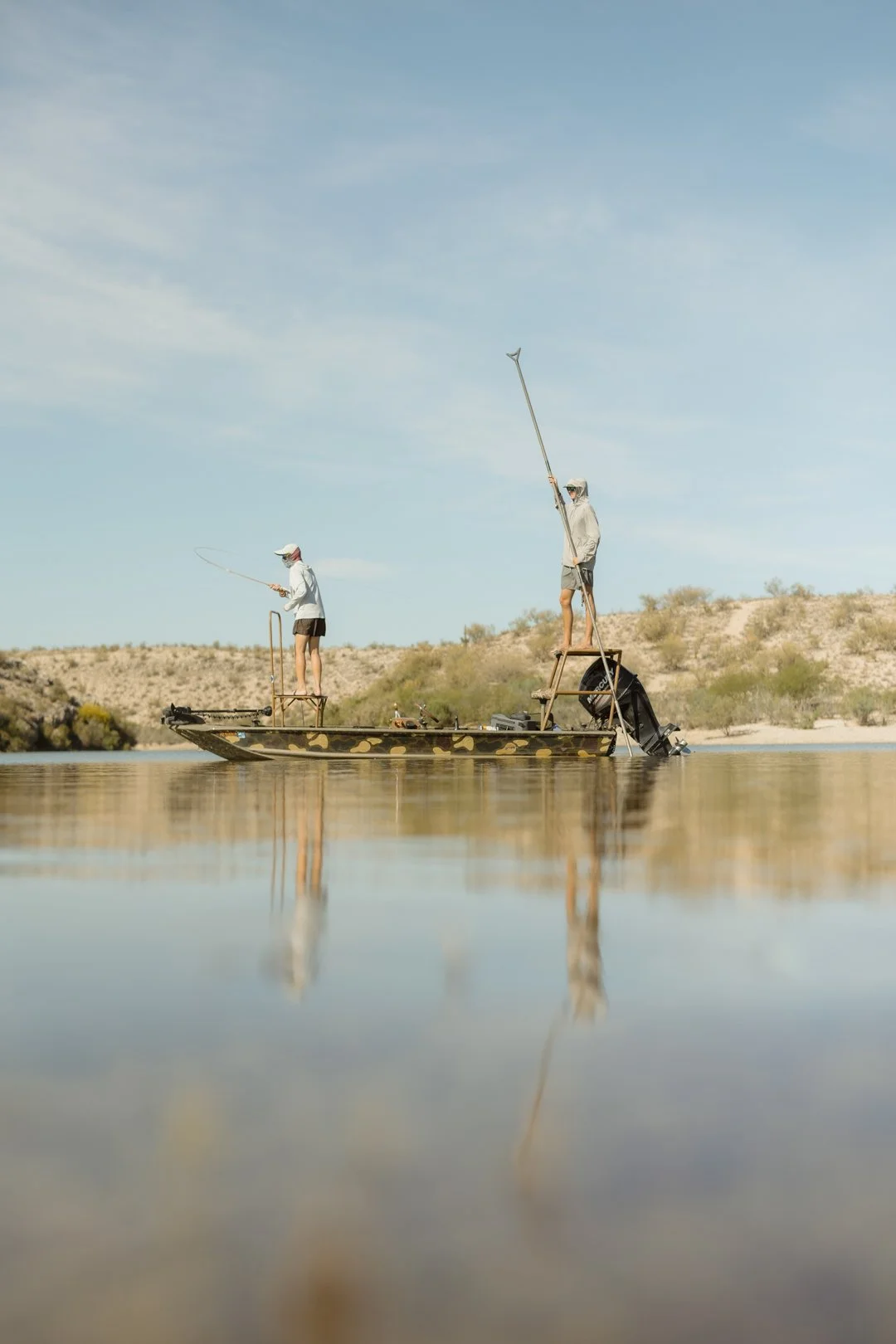 Two people fishing on a boat in a calm body of water, with a desert landscape in the background and clear sky above.