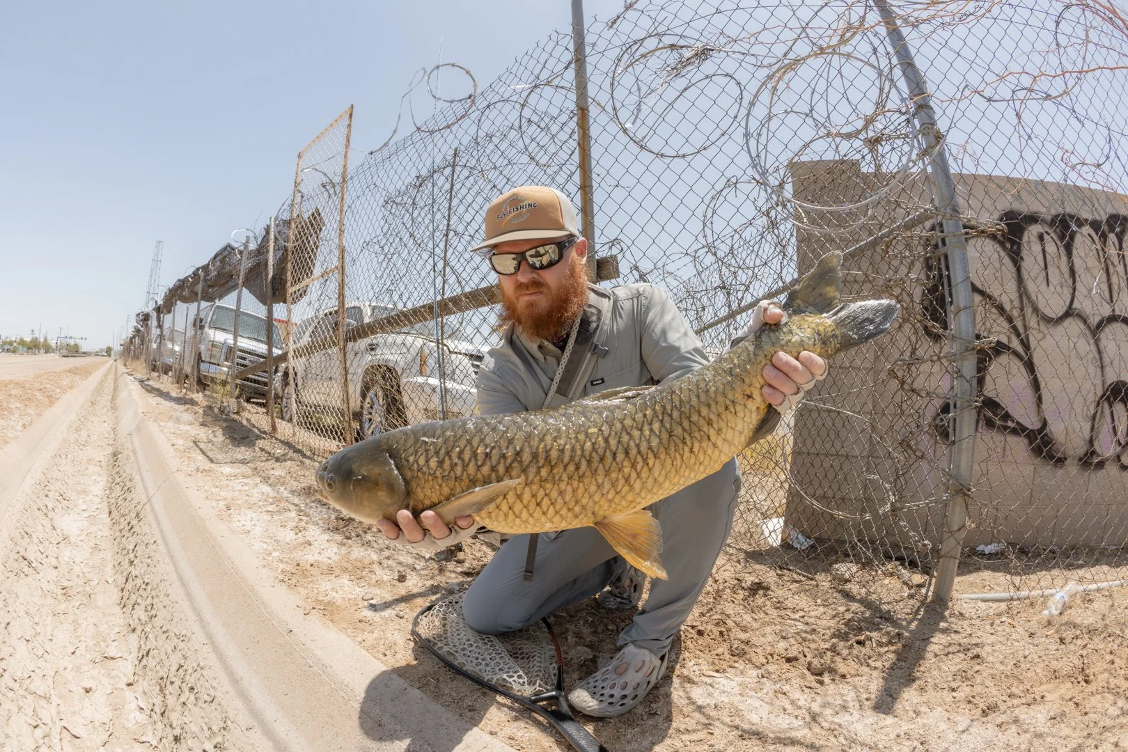 Man with a red beard wearing sunglasses, a beige hat, and a gray outfit kneeling on a dirt road, holding a large fish in front of a chain-link fence with parked cars behind it.