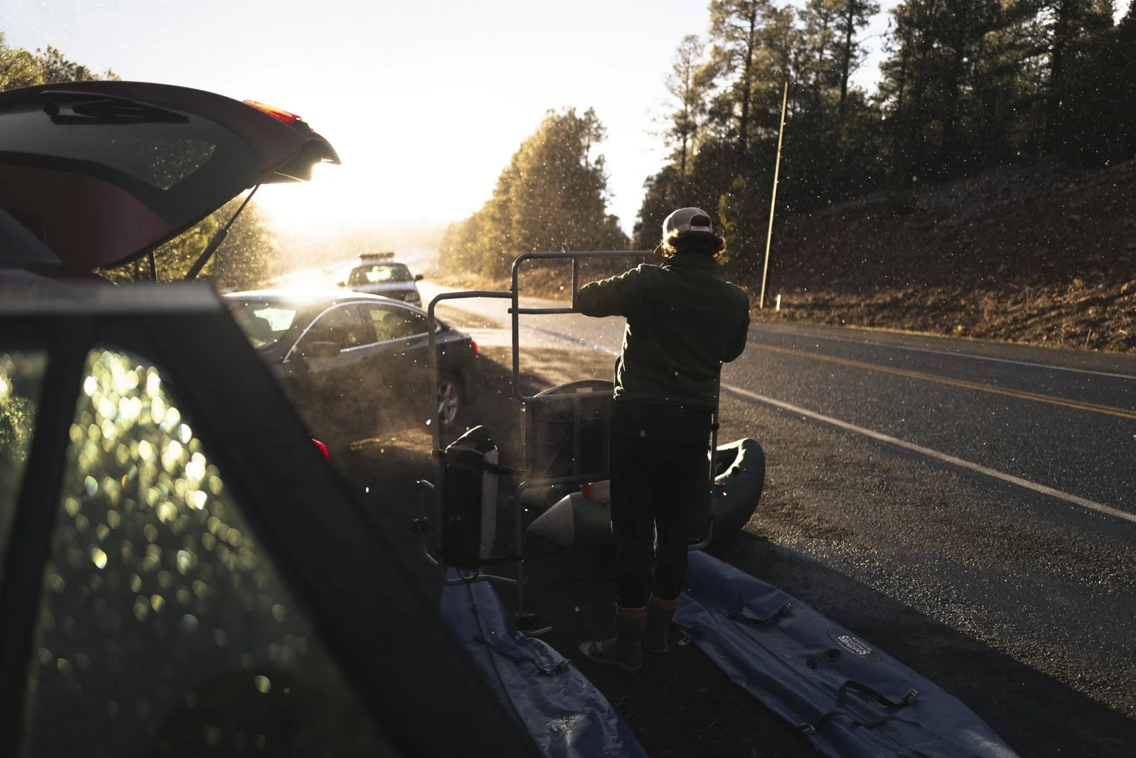 Person on a mobility scooter being assisted by a rescue worker on the side of a two-lane road at sunset, with several parked cars nearby and a wooded hill in the background.