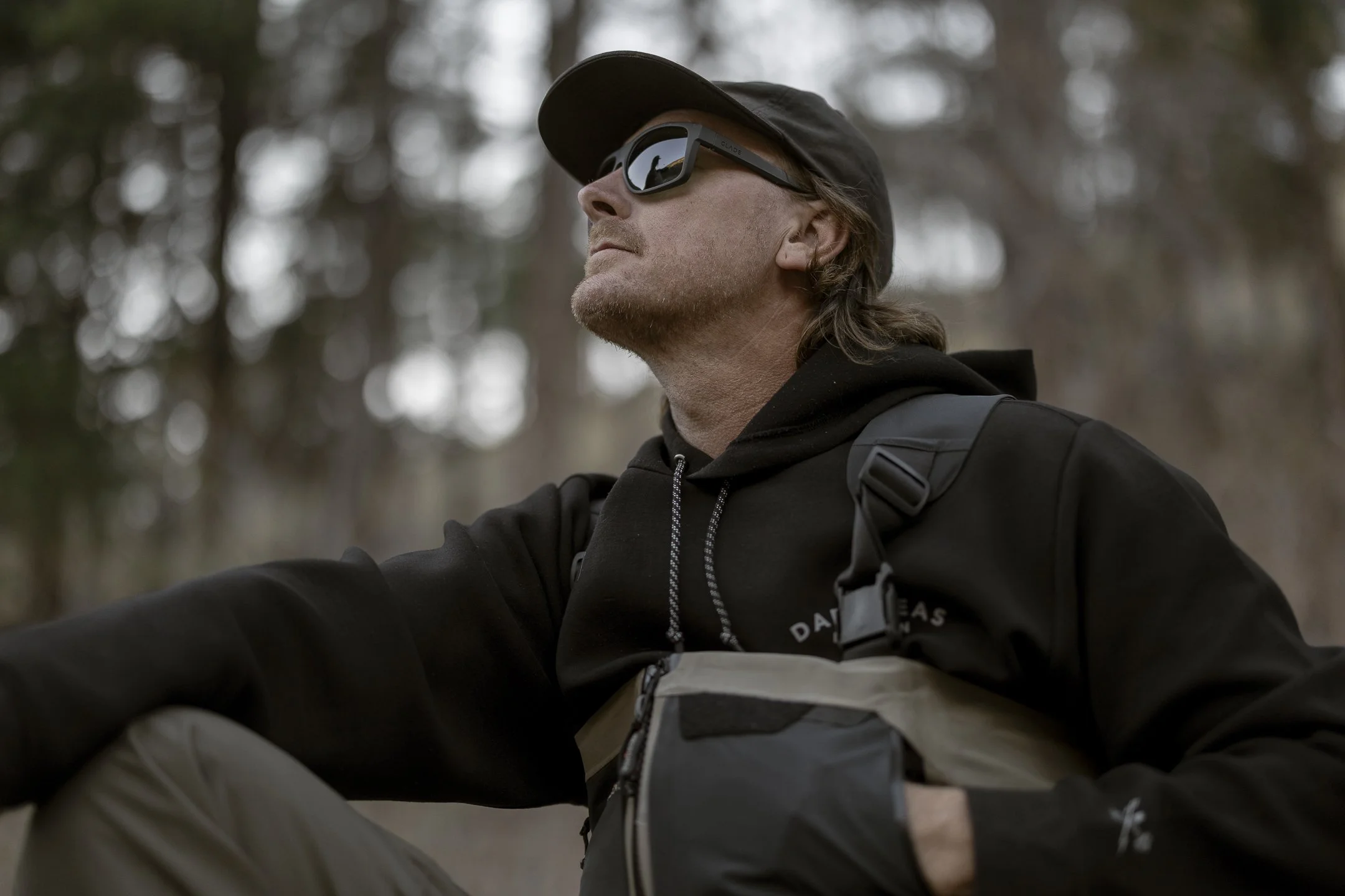A man wearing sunglasses, a black cap, a black hoodie, and a black jacket with a backpack, sitting outdoors in a forest with blurred trees in the background.