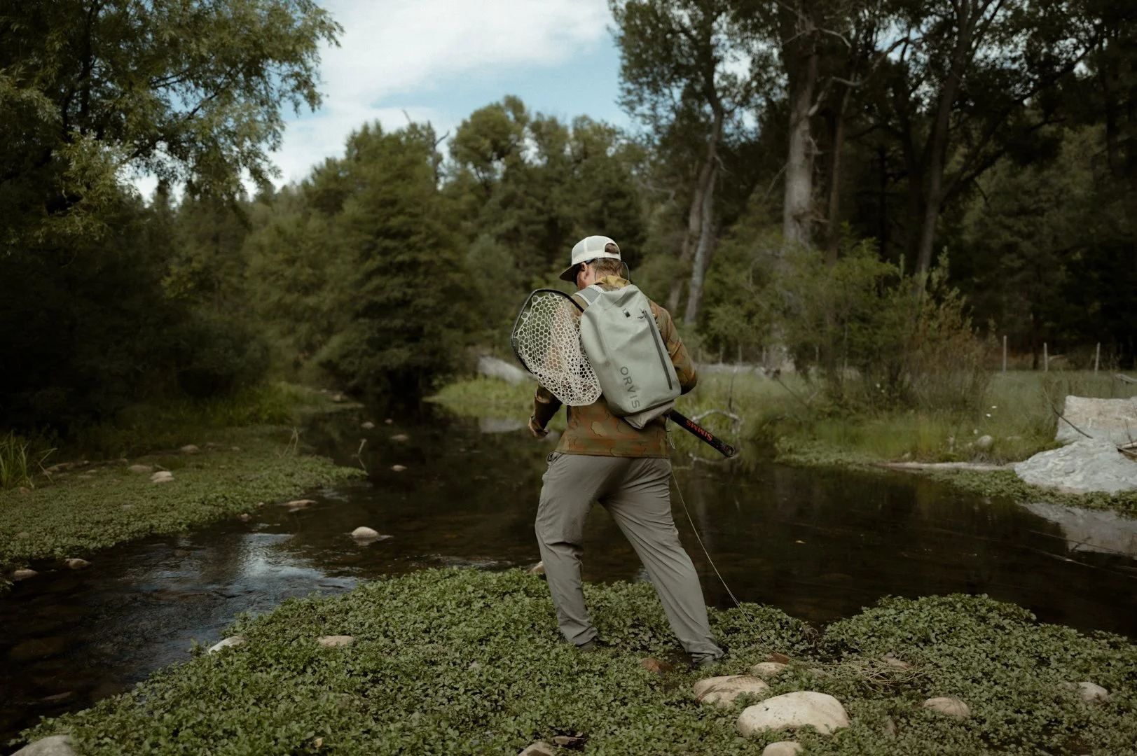 Person with backpack and fishing net standing in a creek surrounded by trees