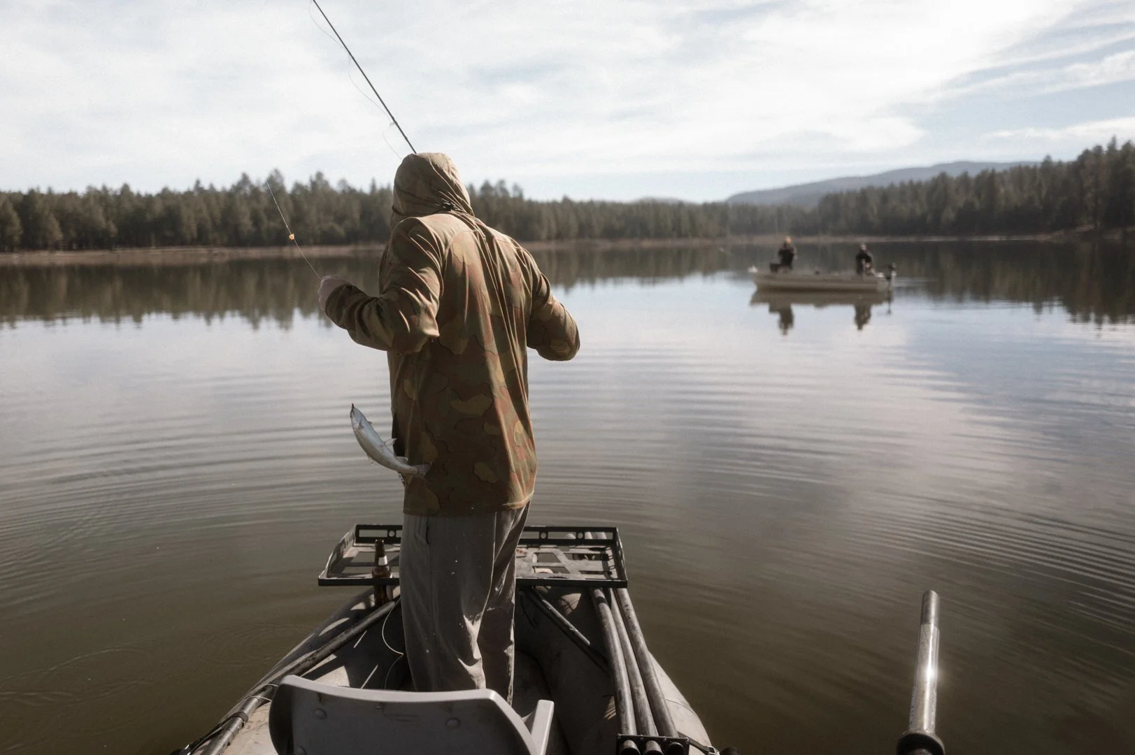 Person fishing in a lake from a boat, with two other people fishing on a boat in the distance, surrounded by trees and mountains under a partly cloudy sky.