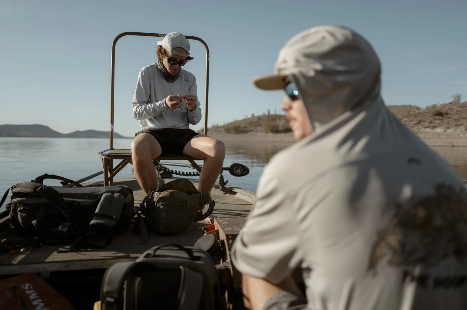 Two men in outdoor gear sitting on a boat, with one looking at a device and the other resting and facing away from the camera, on a calm lake with distant hills.