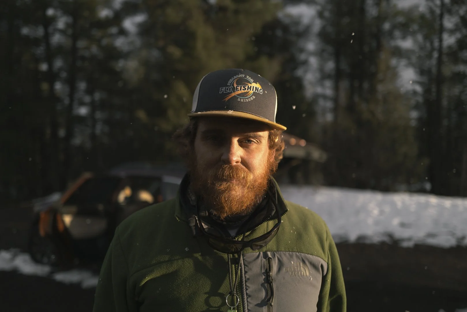 A man with a red beard wearing a black and tan baseball cap that says 'Fly Fishing' and a green North Face jacket, standing outdoors with a snowy forest background.