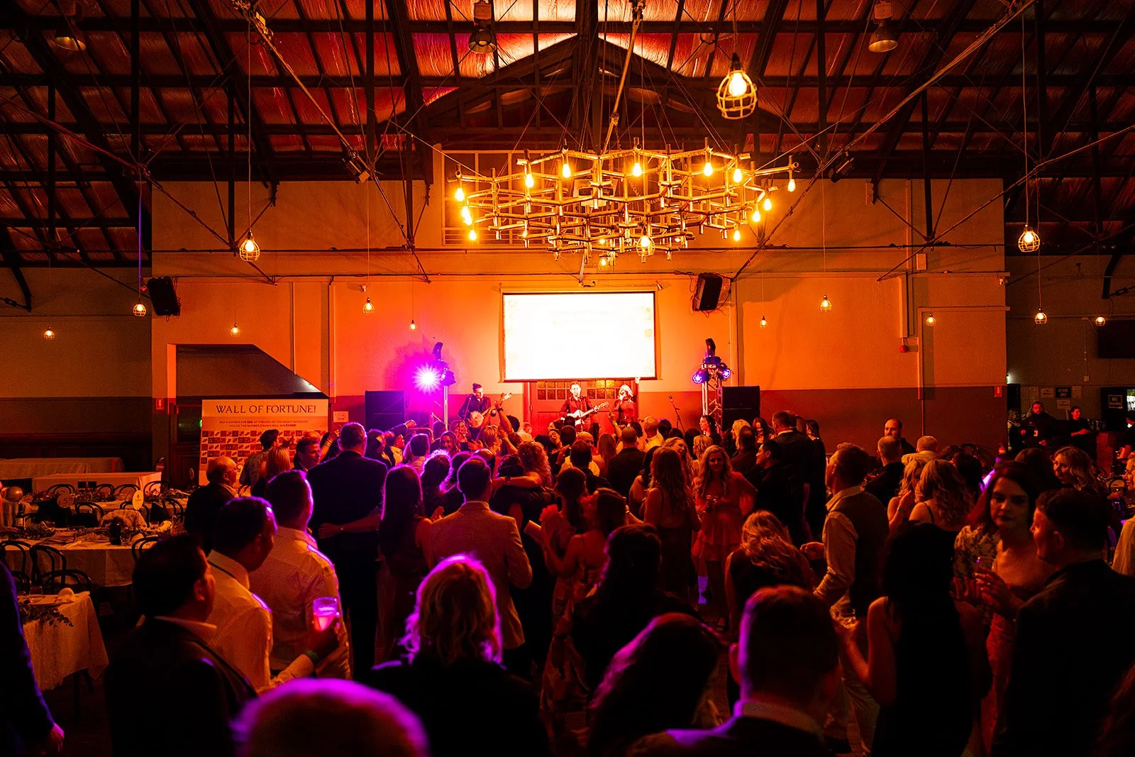 A lively indoor event with a stage where a band is performing, audience dancing and socializing under warm hanging lights in a spacious venue.