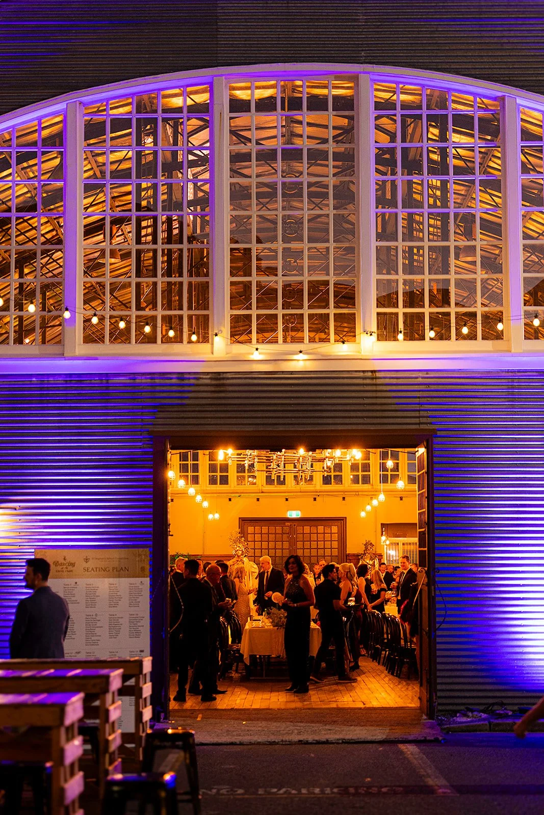People socializing inside a well-lit venue with large windows and purple lighting outside at night.
