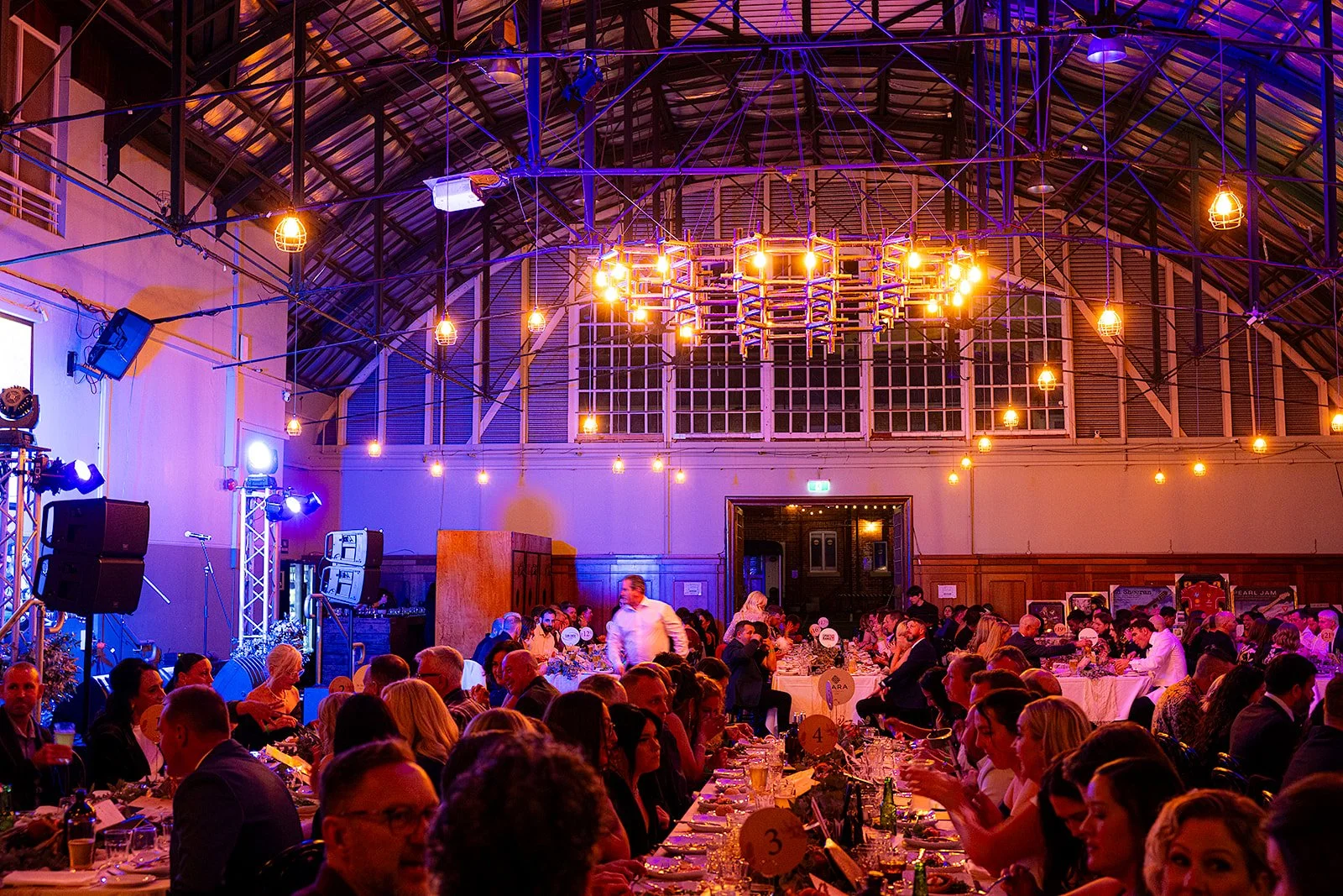 A large banquet hall filled with people sitting at long tables decorated with flowers and tableware. The room has a high, arched ceiling with exposed metal beams and hanging light bulbs. There are purple and orange lighting effects, and a stage with 