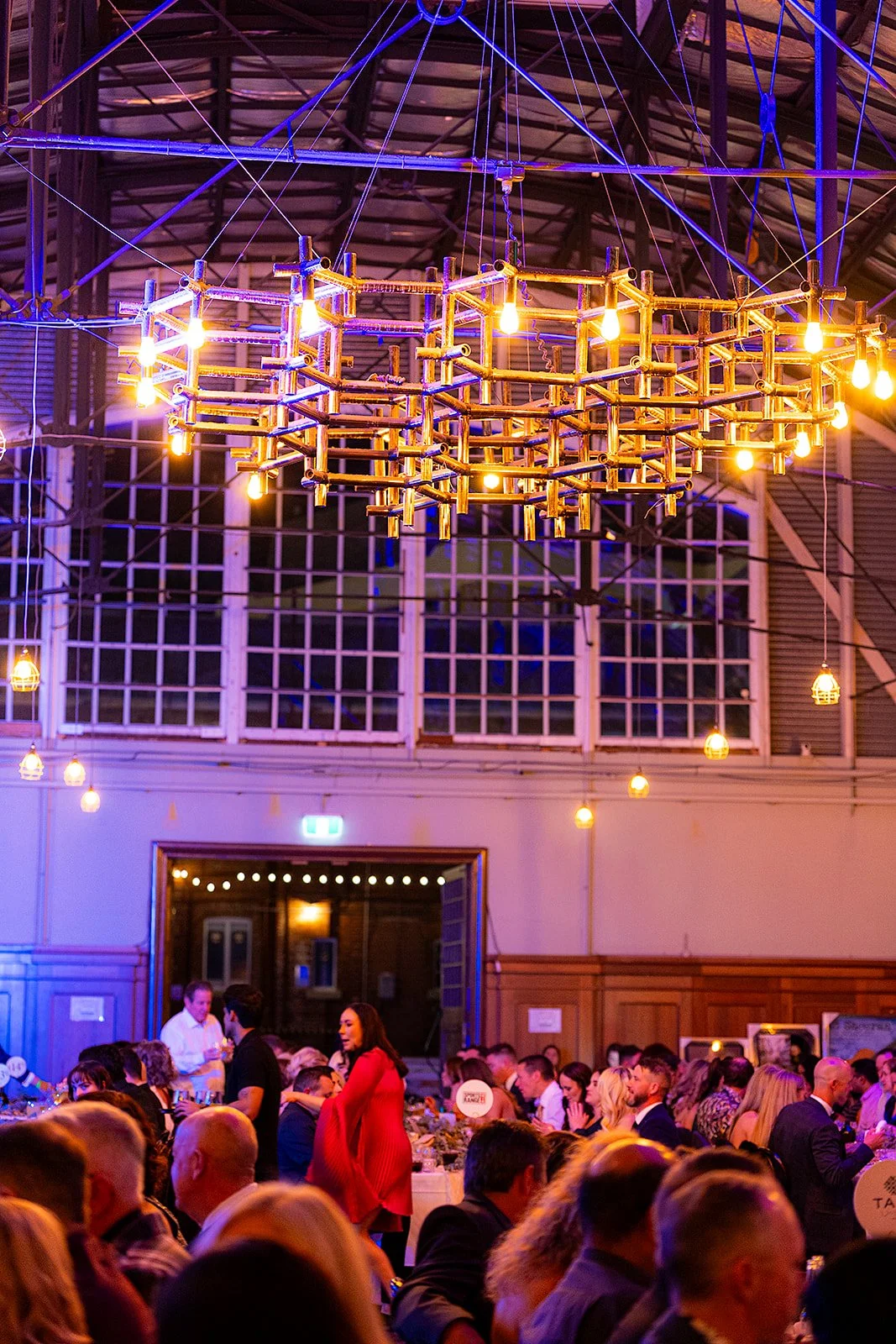 A large chandelier made of interconnected bamboo pipes with exposed light bulbs hangs from the ceiling of a busy indoor event space. The room is filled with many people seated at tables, with some standing and talking, under purple and yellow lightin