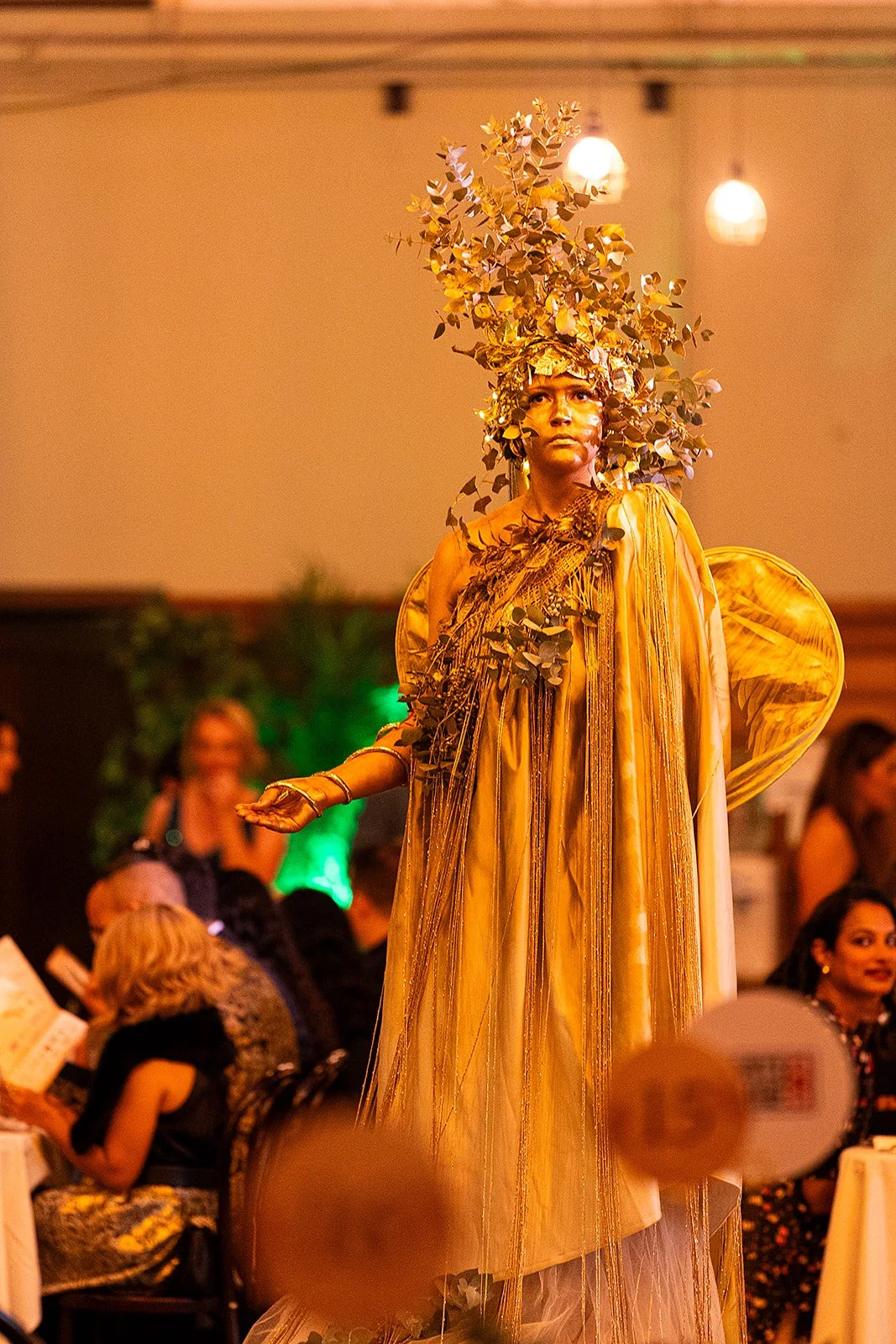 A performer dressed in a gold costume with a headpiece made of metallic leaves and a flowing dress, standing among seated attendees at an event.