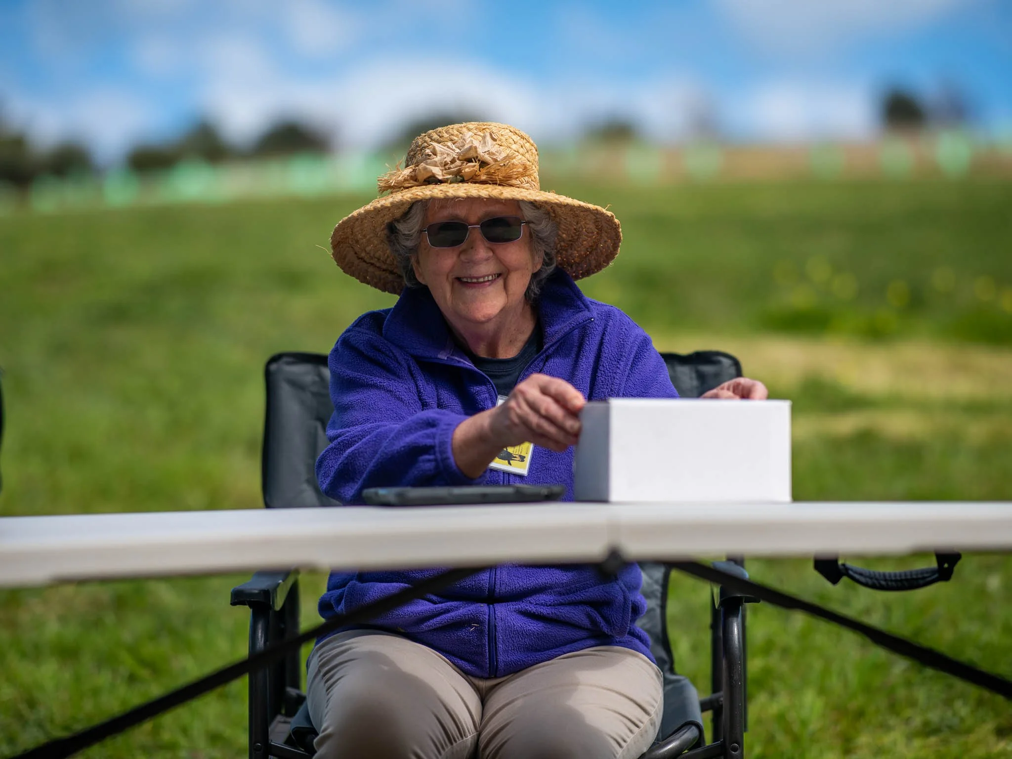 Elderly woman in a straw hat and sunglasses sitting outdoors at a table with a white box, smiling, with a grassy background.