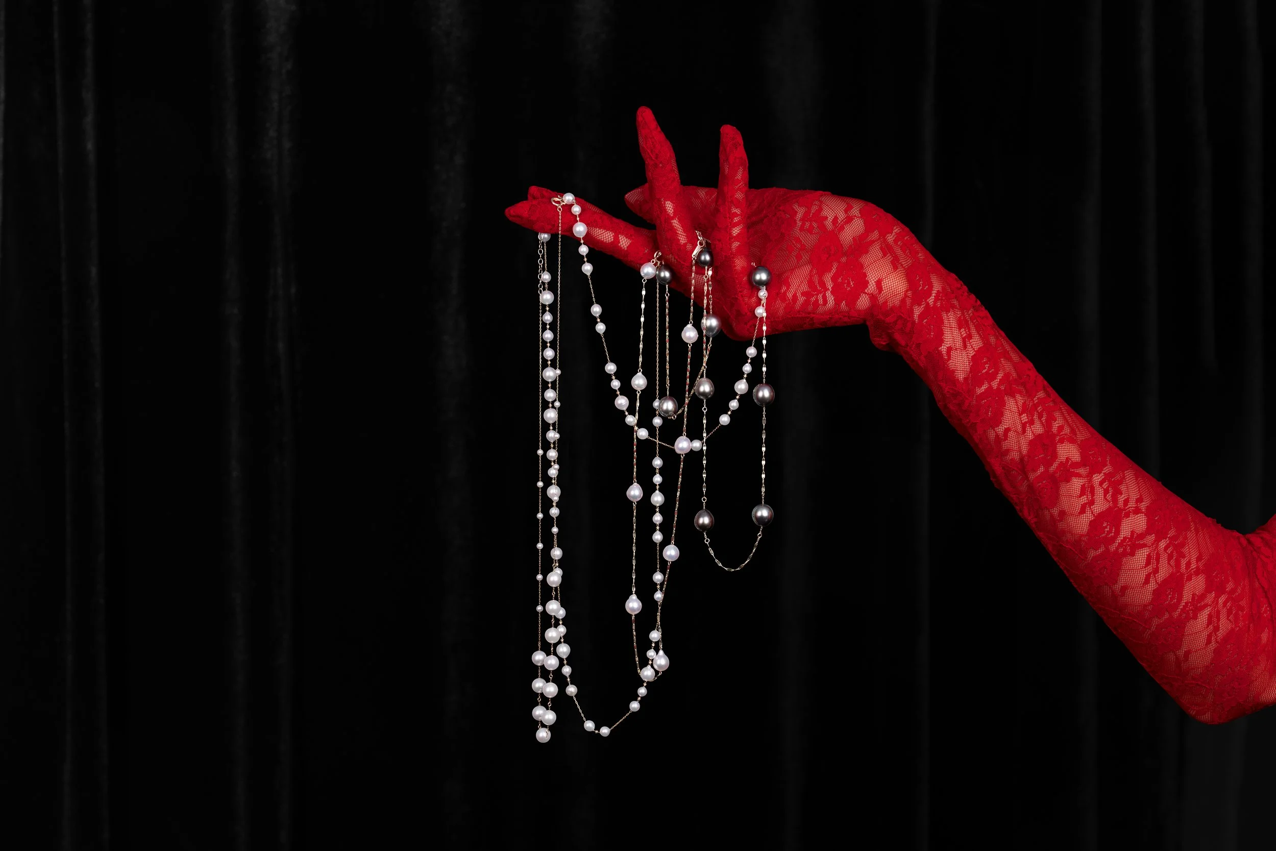A hand in a red lace glove holding and displaying multiple strands of pearl and silver necklaces against a black curtain background.