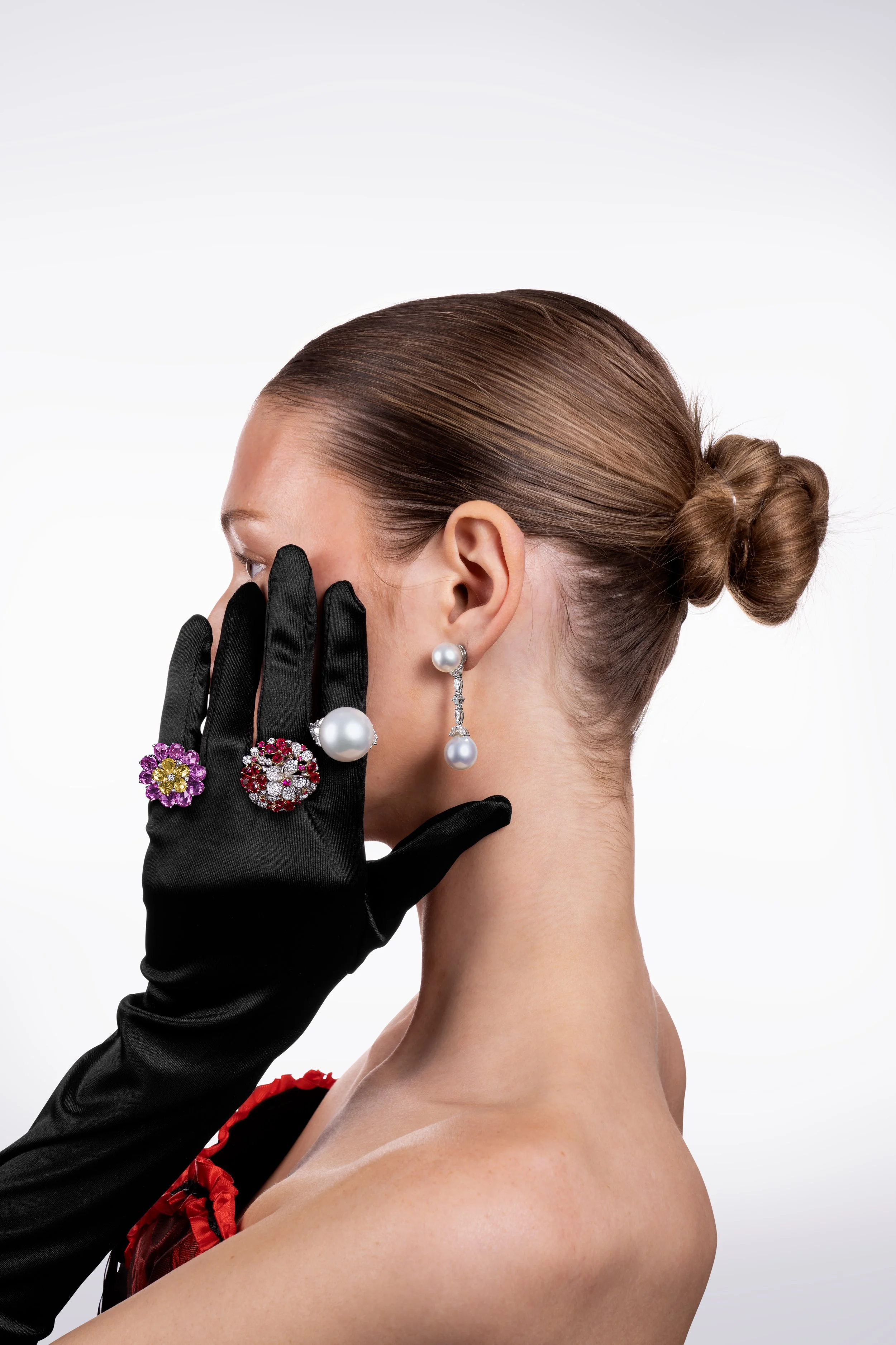 A woman with brown hair styled in a bun, wearing pearl earrings, black gloves, and a dress with a red and black floral design. She is touching her face with her gloved hand, which has multiple ornate rings with large decorative stones. The background