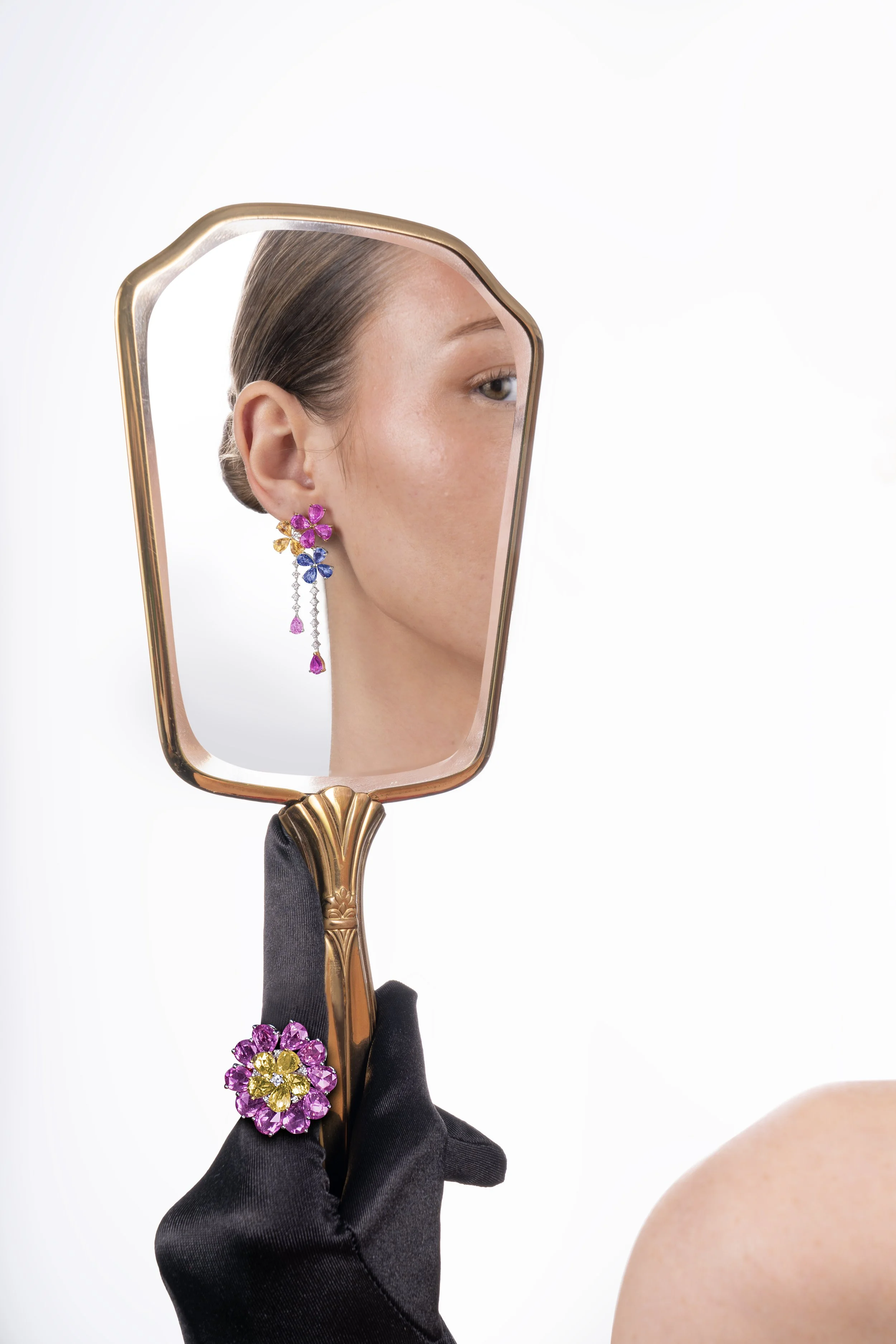 Close-up of a woman looking into a hand mirror, showing her face with a focus on her cheek and earring. She has smooth skin, well-groomed hair, and is wearing pink, yellow, and blue floral earrings with a dangling chain.
