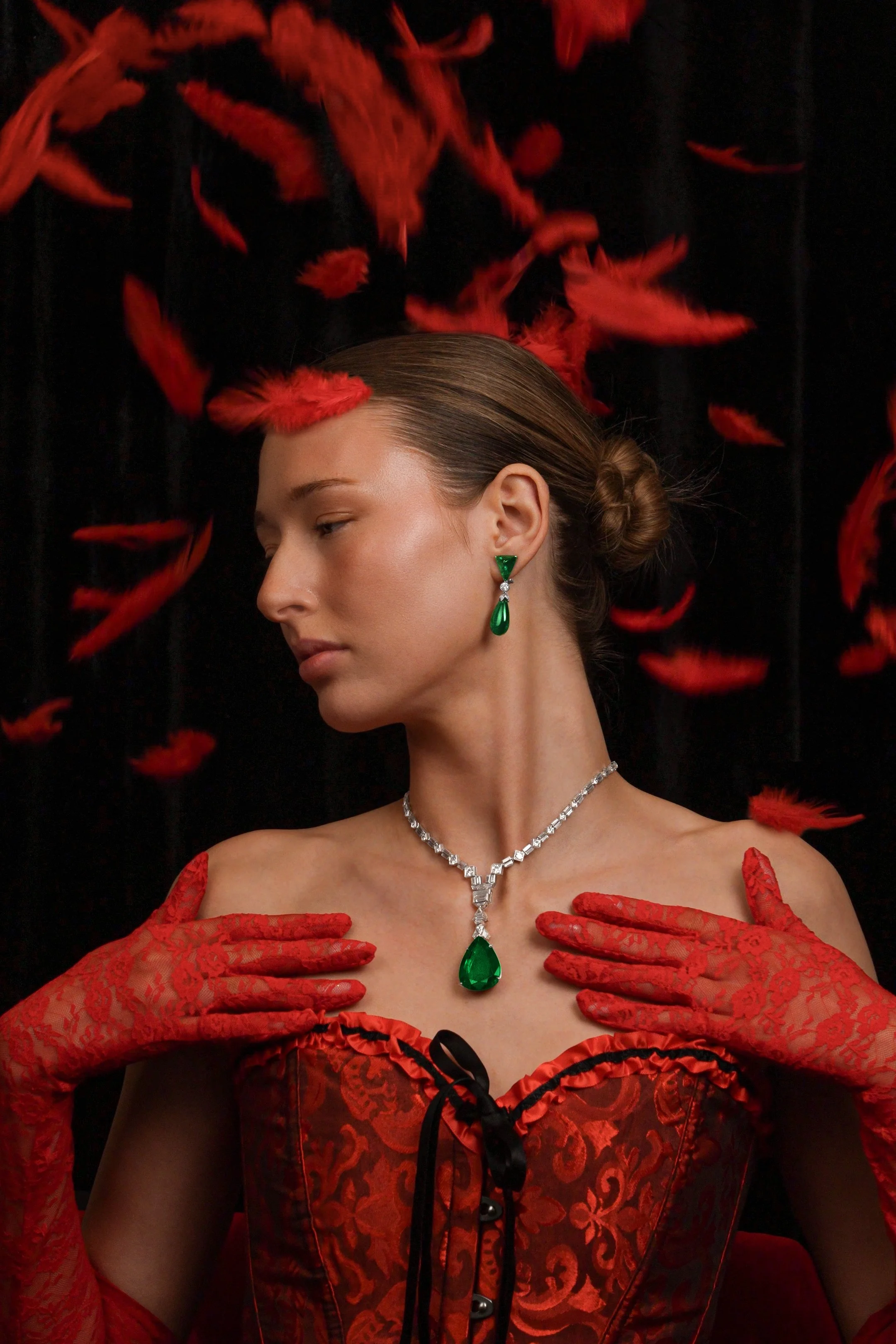 A woman dressed in a red lace gown with black accents and red lace gloves, wearing Emeralds while feathers are falling around her. 
