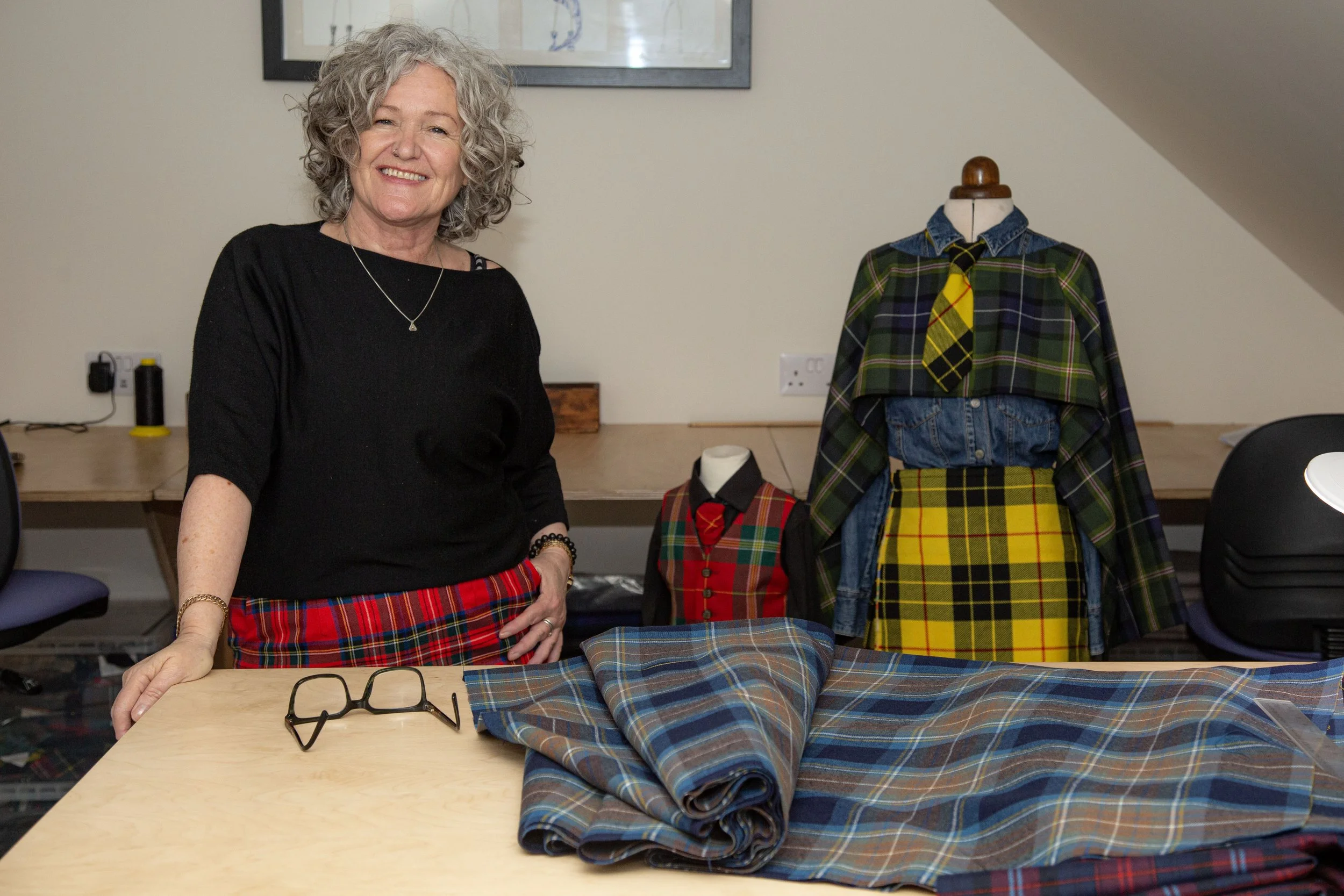 Grey Haired lady wearing red tartan trousers, standing by a desk with dummies clothe in tartan beside her