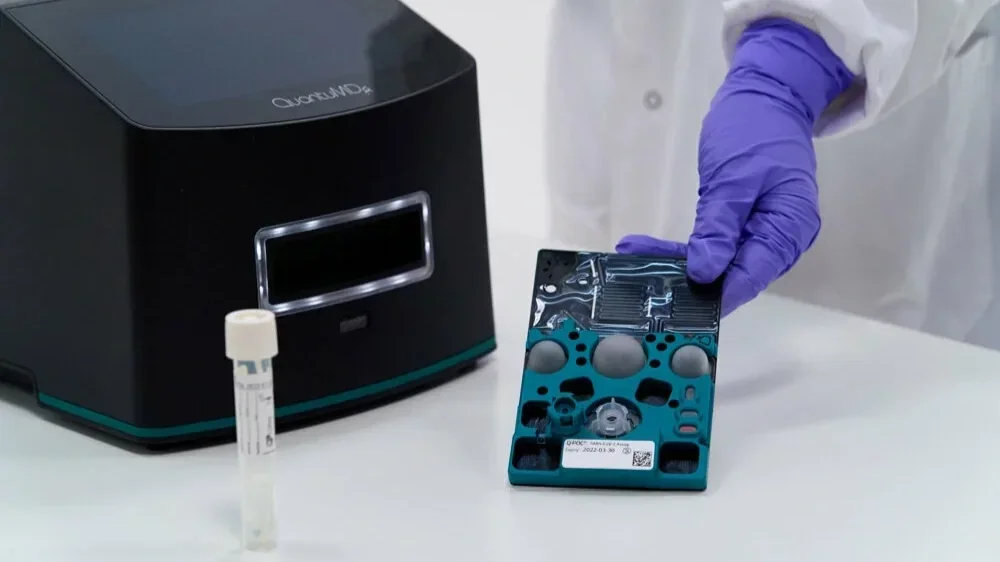 QuantuMDx Q-POC system including an instrument reader, a sample tube and a Q-POC PCR test cartridge held by a lab technician wearing purple gloves
