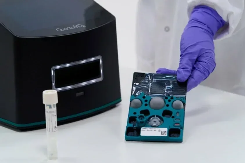 QUantuMDx Q-POC cartridge held by a lab technician with purple gloves, Q-POC instrument and a sample test tube