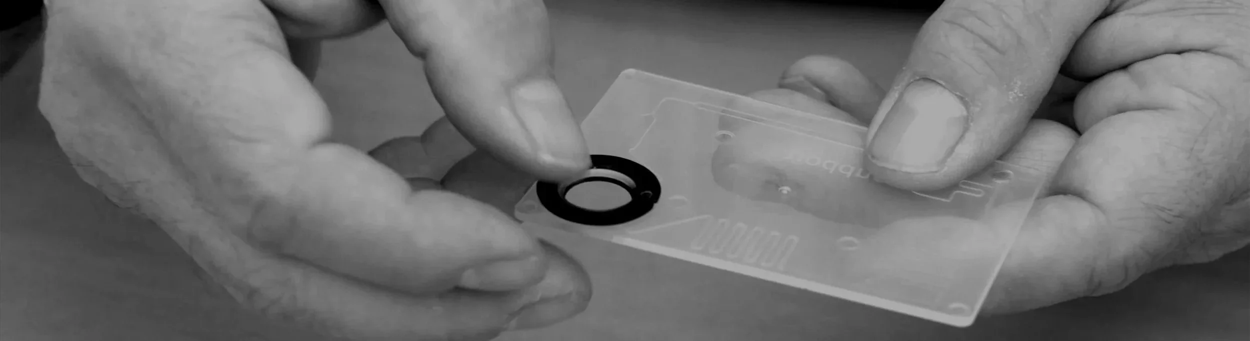 Close-up of a person placing a black circular PumpRing in a transparent plastic microfluidic cartridge.