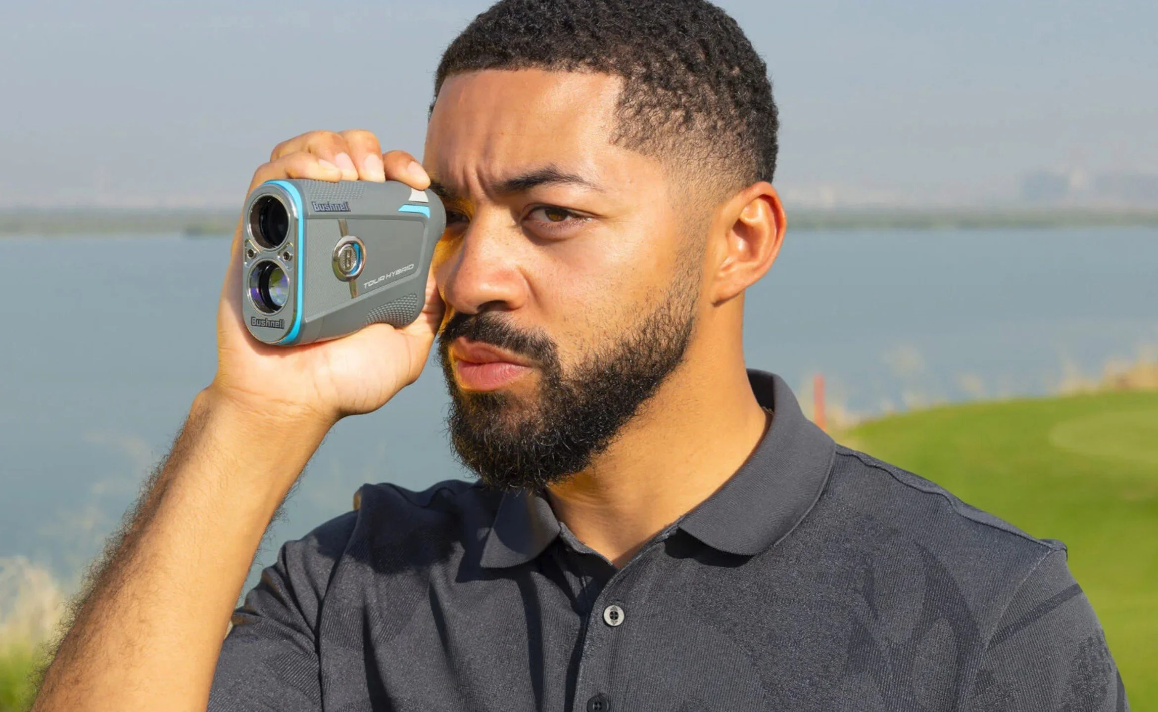 A man with a beard wearing a gray polo shirt is holding a small camera to his eye, taking a photo outdoors with a body of water and green landscape in the background.