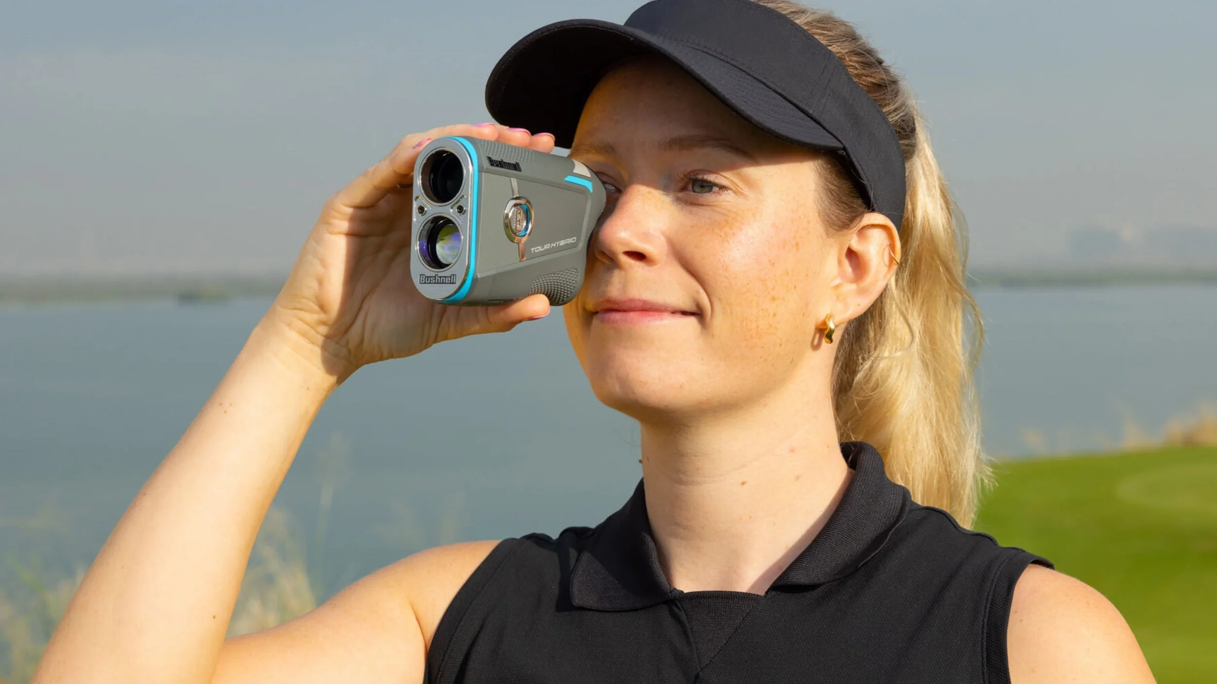 Woman with blonde hair, wearing a black visor and sleeveless black sports top, looking through a small digital camera while outdoors near a body of water.