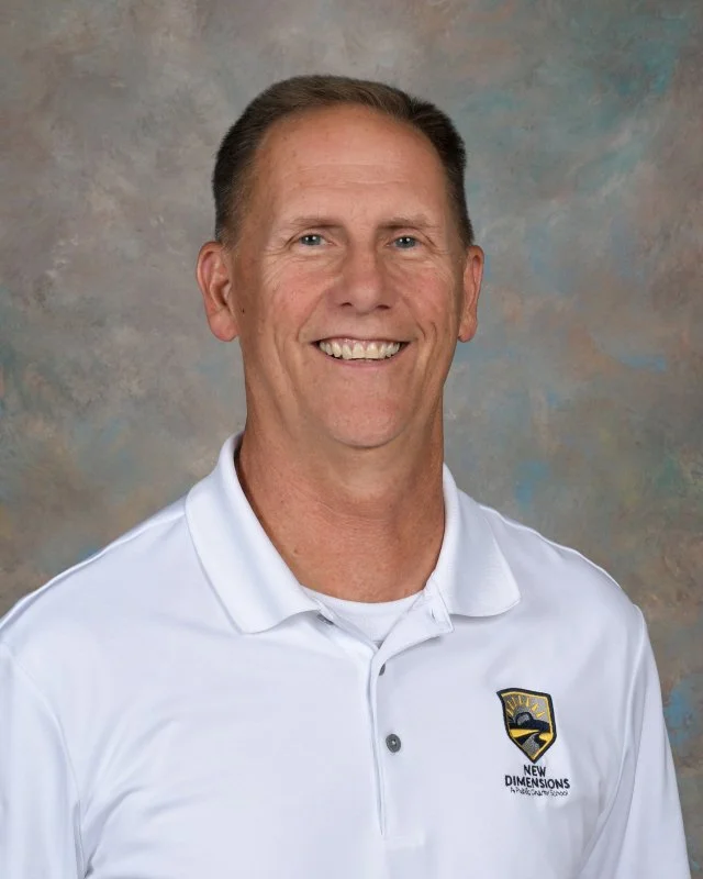 Portrait of a man smiling, wearing a white polo shirt with a logo that reads 'New Dimensions' and features a stylized image of the sun and a road.