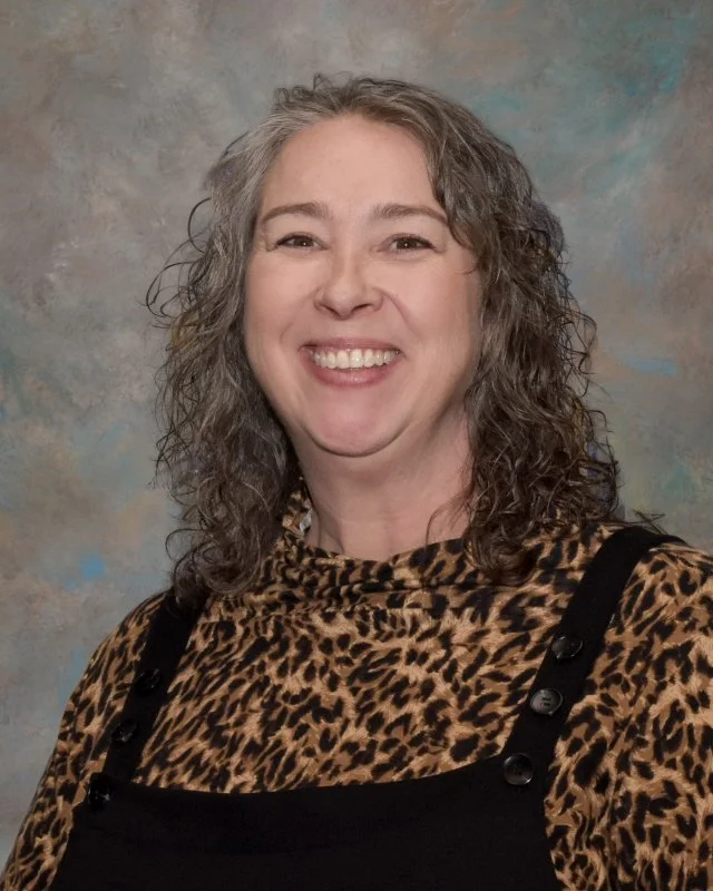 A woman with curly gray hair smiling, wearing a leopard print top and black overalls, in front of a textured background.