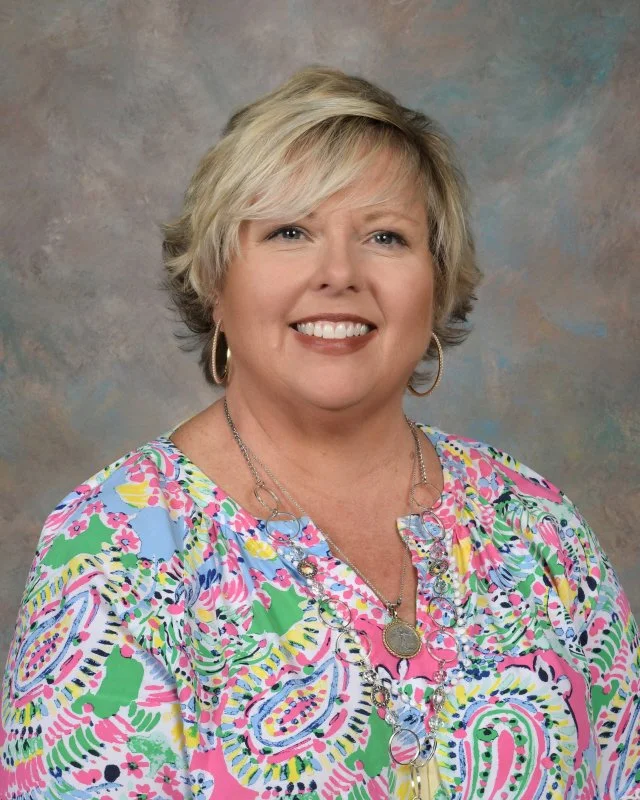 A woman with short, blonde hair smiling at the camera, wearing a pink paisley top, against a neutral textured background.