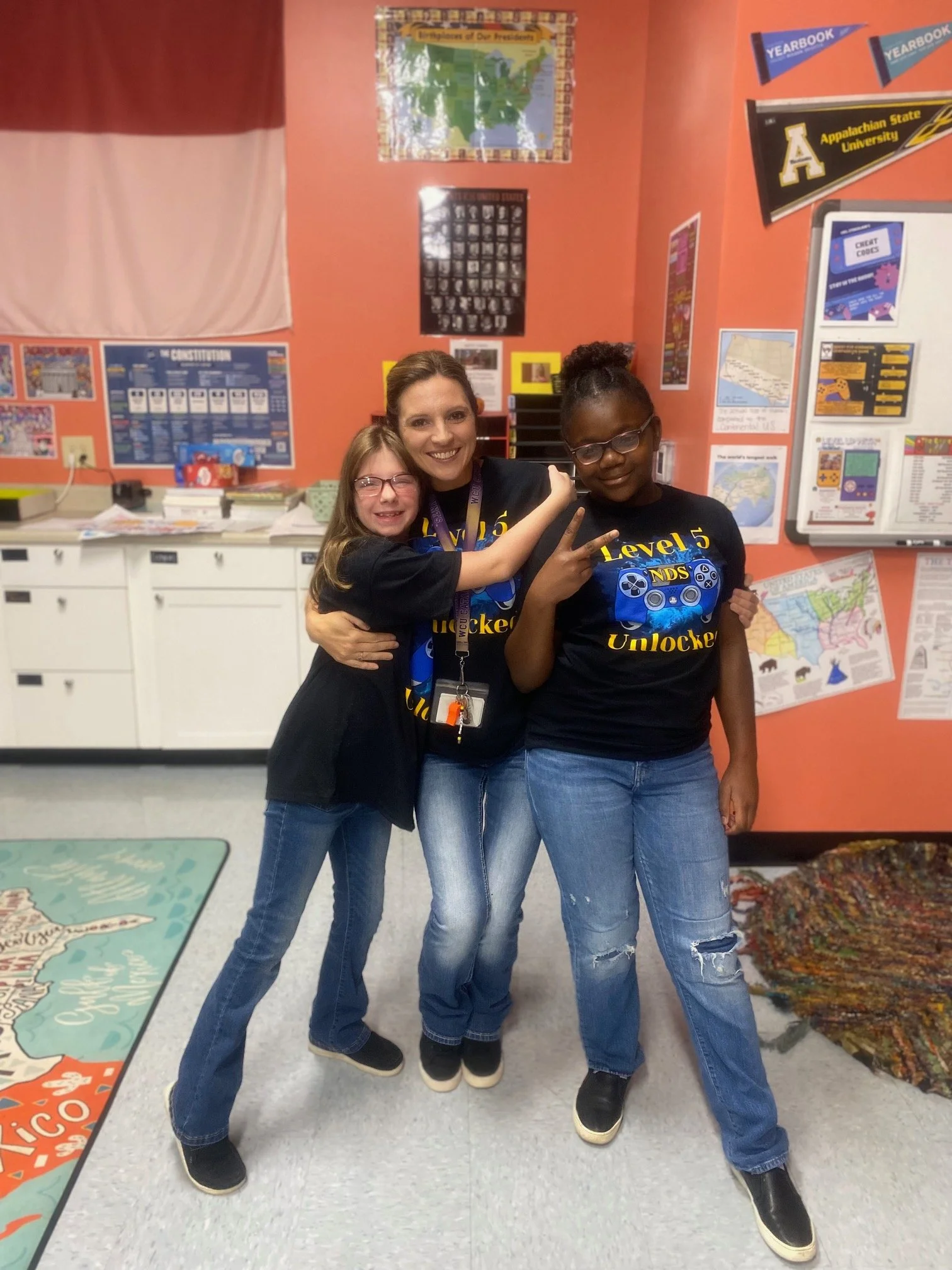 Three young girls and a woman standing together in a classroom, smiling and making peace signs. The classroom walls are decorated with posters, maps, and school banners.
