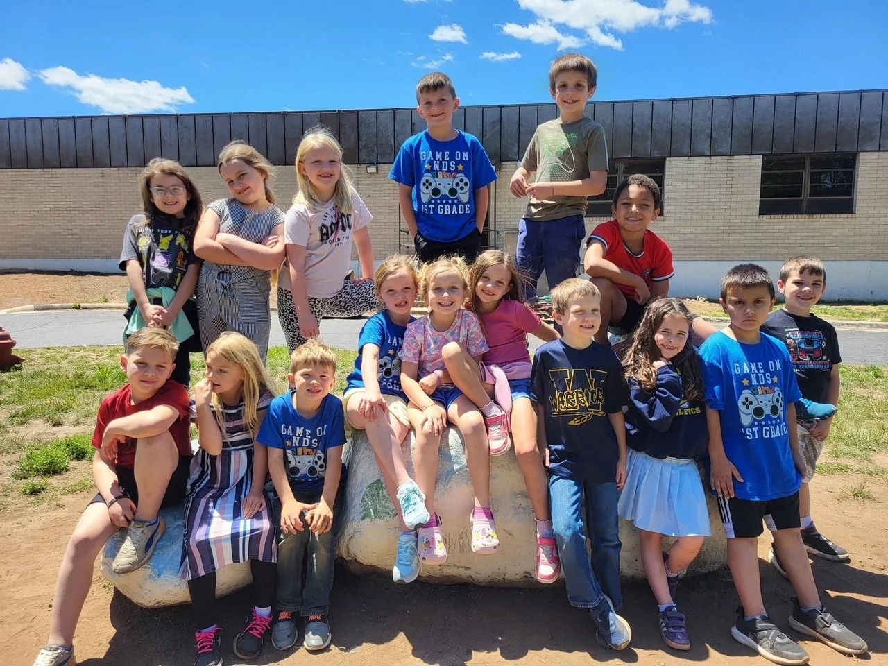 Group of young children posing together outdoors on a sunny day, with some sitting and others standing in front of a school building.