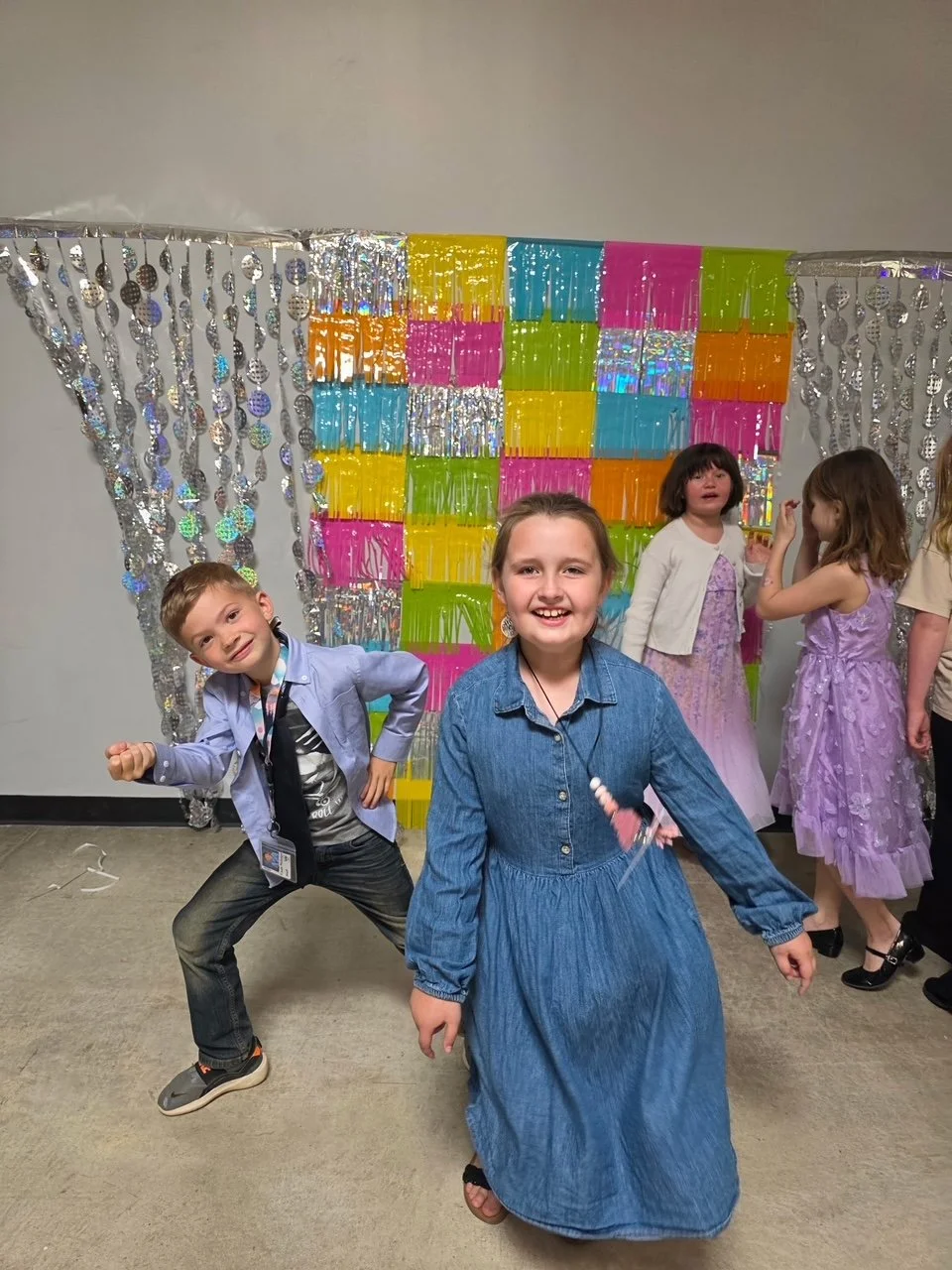 Children at a festive event, with colorful backdrop of shiny decorations and gift boxes, smiling and posing for the camera.