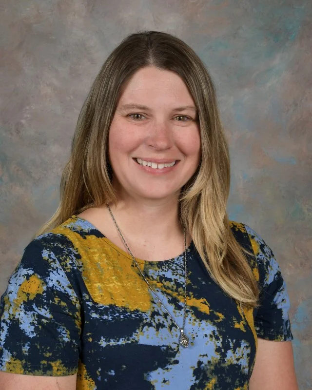 A woman with long, light brown hair smiling at the camera, wearing a colorful, abstract patterned top in blue, yellow, and black with a pendant necklace, against a neutral textured background.