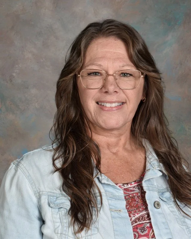 A woman with long wavy brown hair, wearing glasses, a denim jacket, and a patterned red and white top, smiling at the camera against a mottled background.
