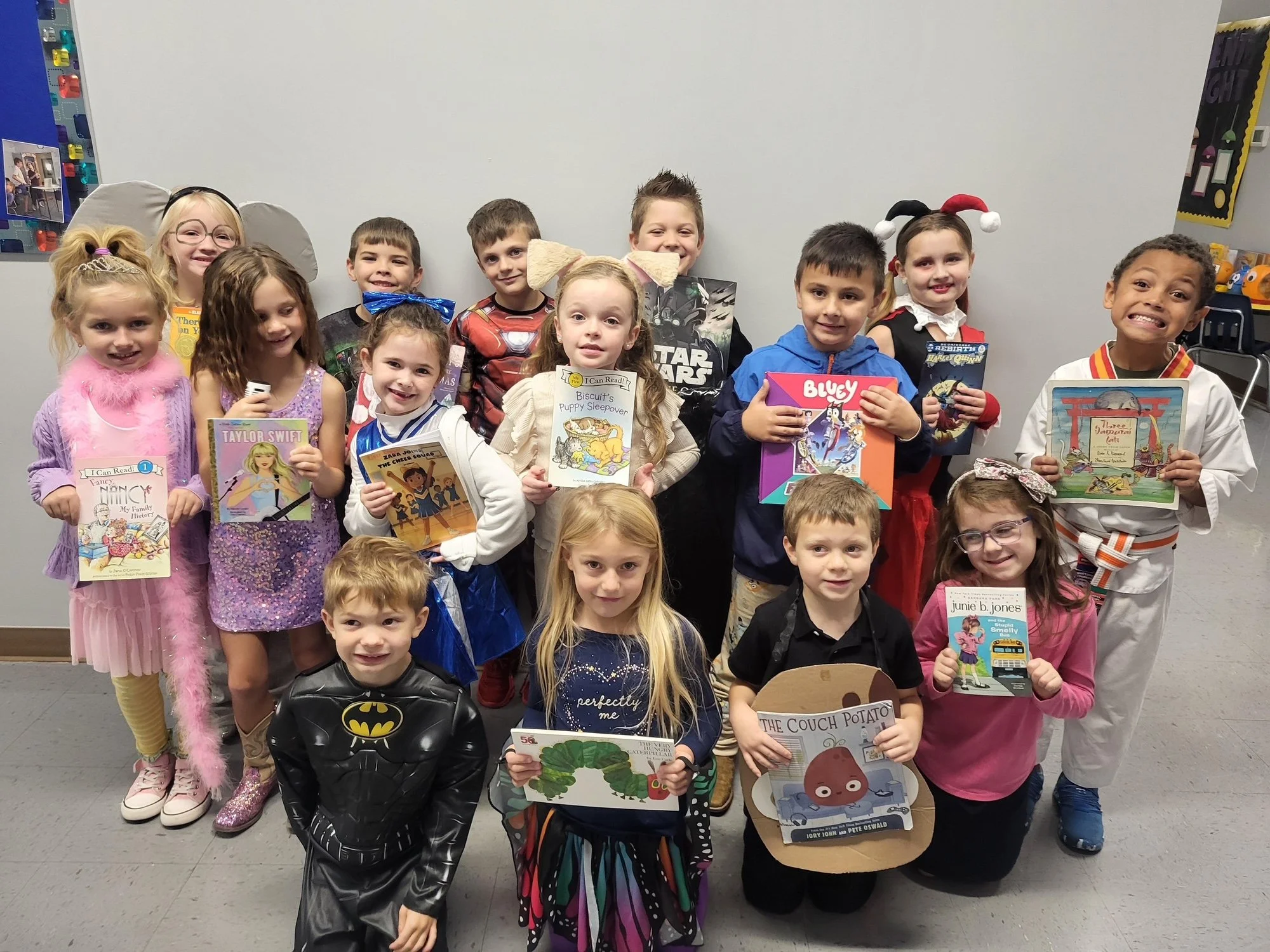 Group of children dressed in costumes and holding books, smiling indoors against a plain white wall.