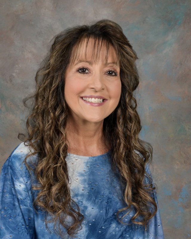 A woman with long, curly brown hair and a warm smile, wearing a blue tie-dye top, against a neutral, textured background.