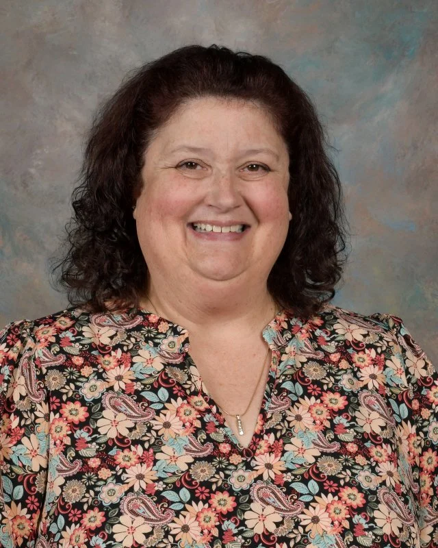 A woman with curly dark hair smiling, wearing a floral blouse with various pink, red, blue, and beige flowers and paisley patterns, against a gray textured background.