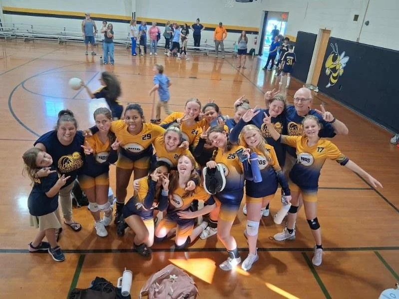 A group of young girls and their coach celebrating on a gymnasium floor after a volleyball game, dressed in yellow and blue uniforms, some making peace signs, with other people in the background near the gym walls.
