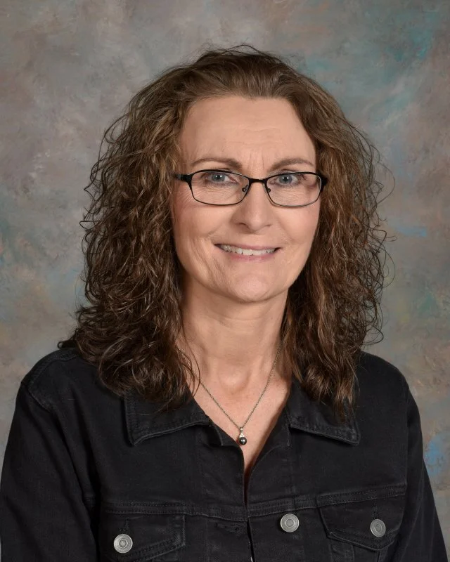A middle-aged woman with curly brown hair and glasses, wearing a black denim jacket and a silver necklace, smiling in front of a textured background.