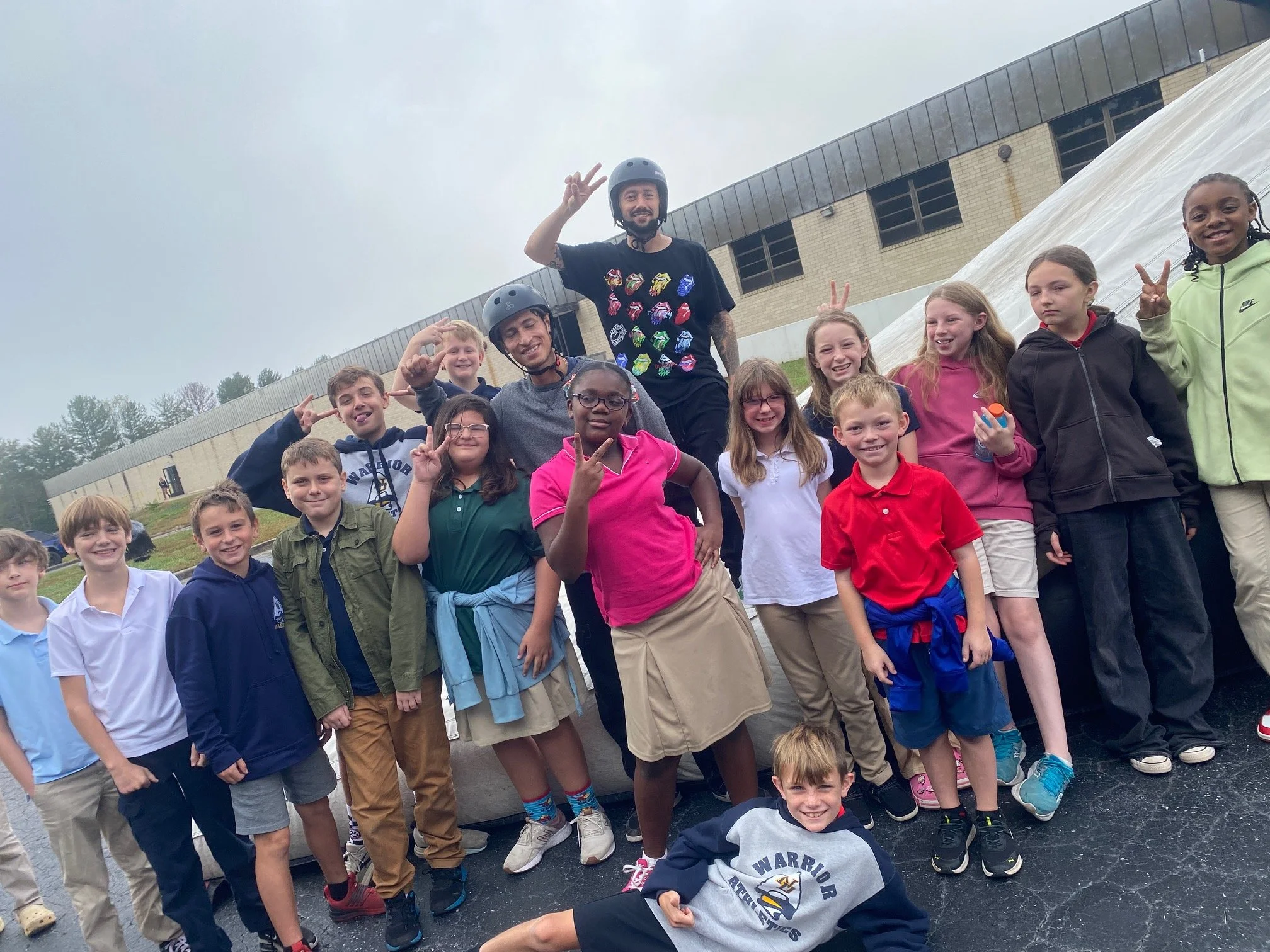 A group of children and two adults wearing helmets and casual clothes, standing outdoors in front of a building with a sloped roof and surrounded by trees. The group is smiling, making peace signs, and enjoying an outdoor activity.
