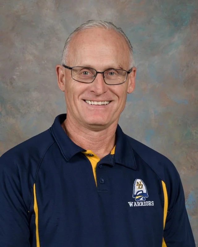 A middle-aged man wearing glasses and a navy blue sports polo shirt with yellow accents, smiling, in front of a multicolored studio background.
