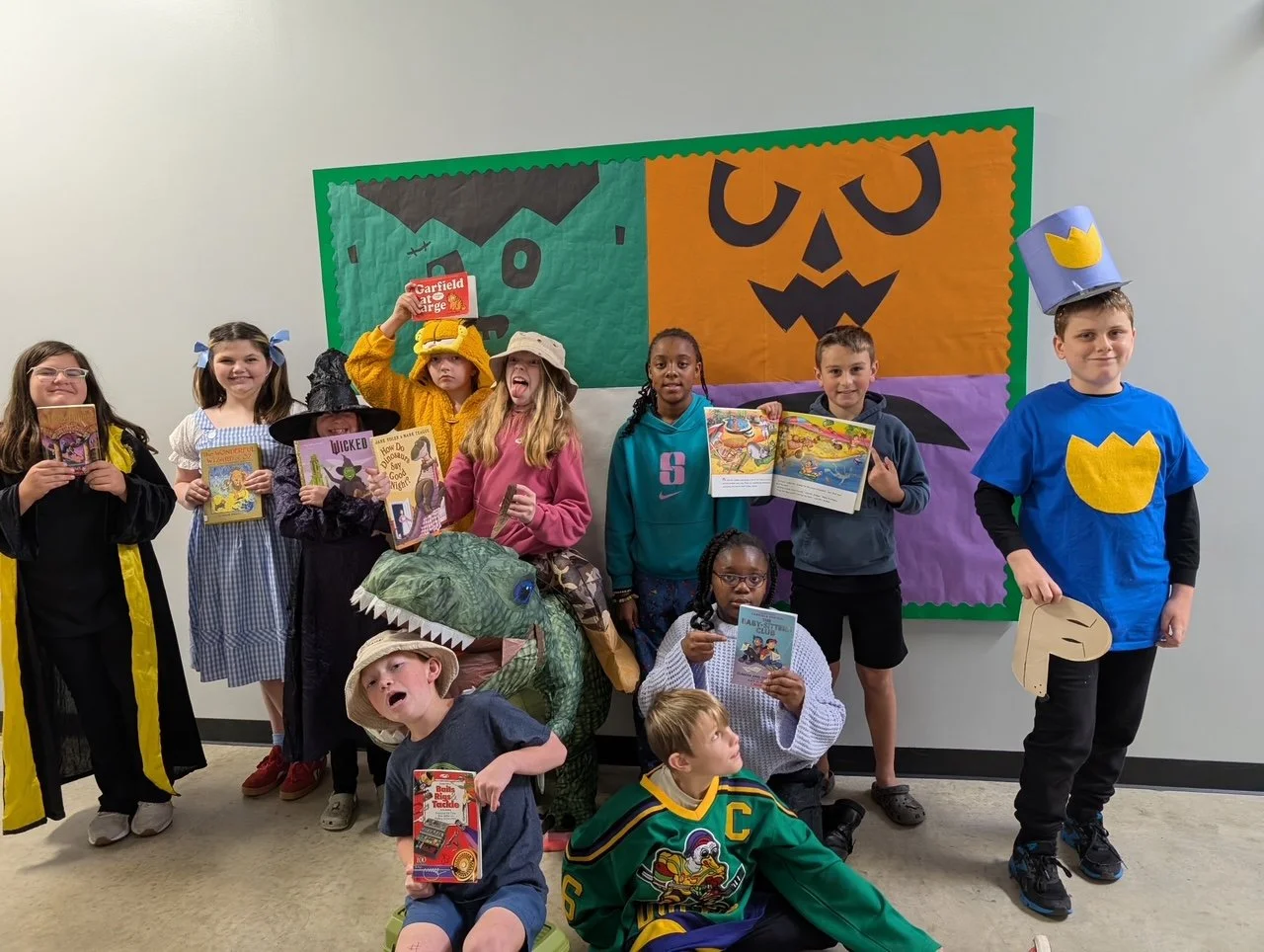 Group of children dressed in costumes, some holding books, posing in front of a Halloween-themed bulletin board decorated with a pumpkin face.