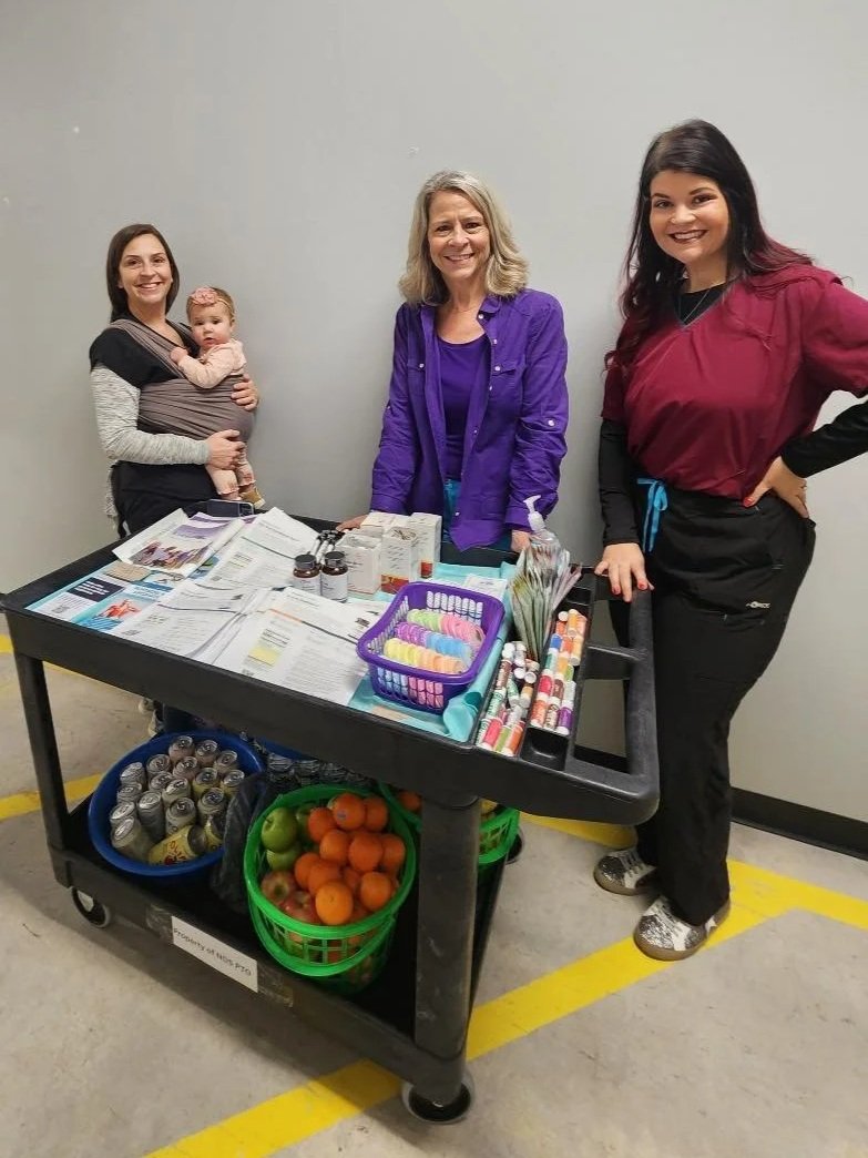 Three women and a baby standing behind a black cart filled with medical supplies, fruit, and canned drinks in a warehouse or storage area.